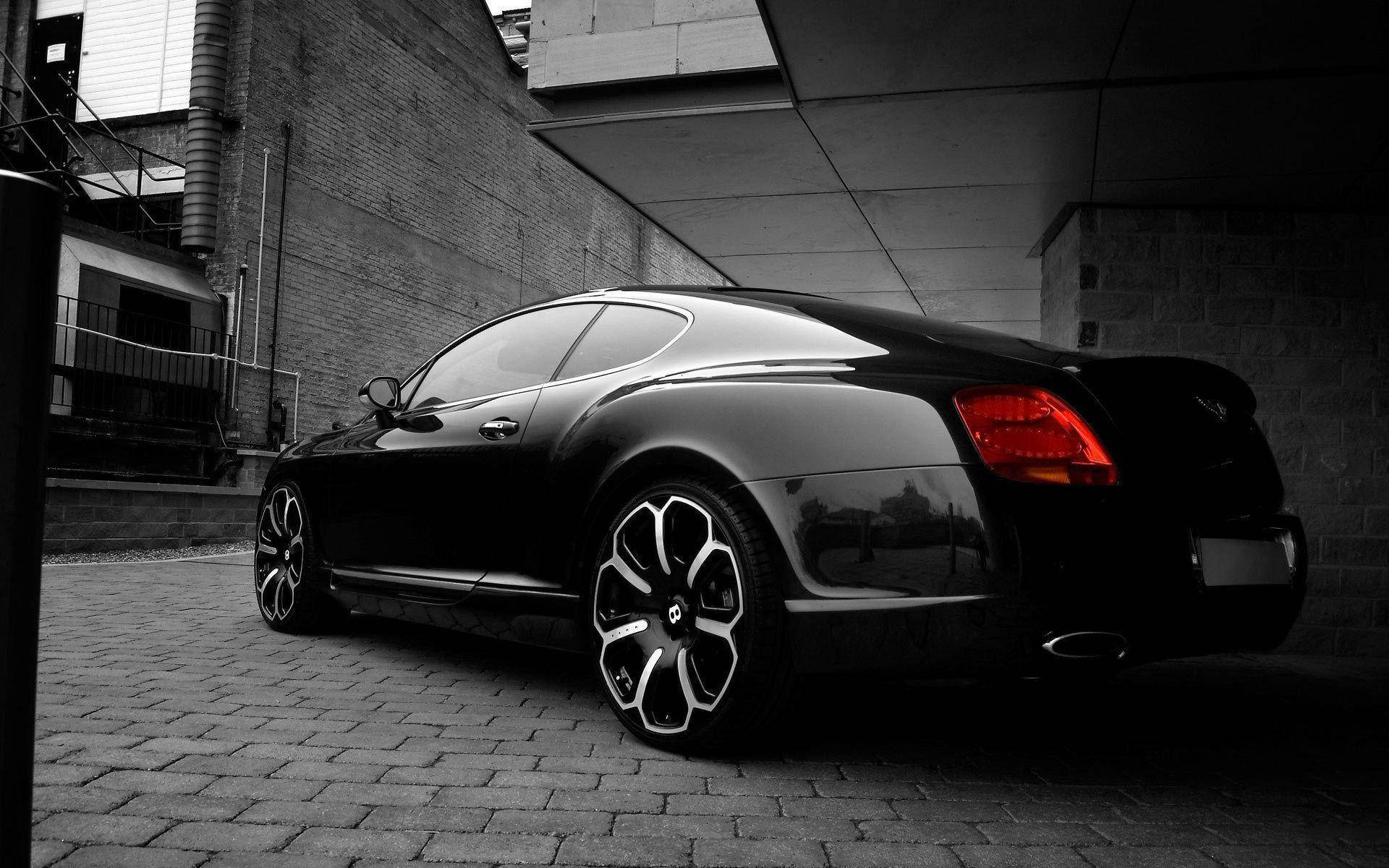 bentley HD 桌布 壁紙 — 一輛時尚的黑色賓利 Continental GT 停泊在磚牆與混凝土建築之間的鵝卵石小巷，光影對比強烈。