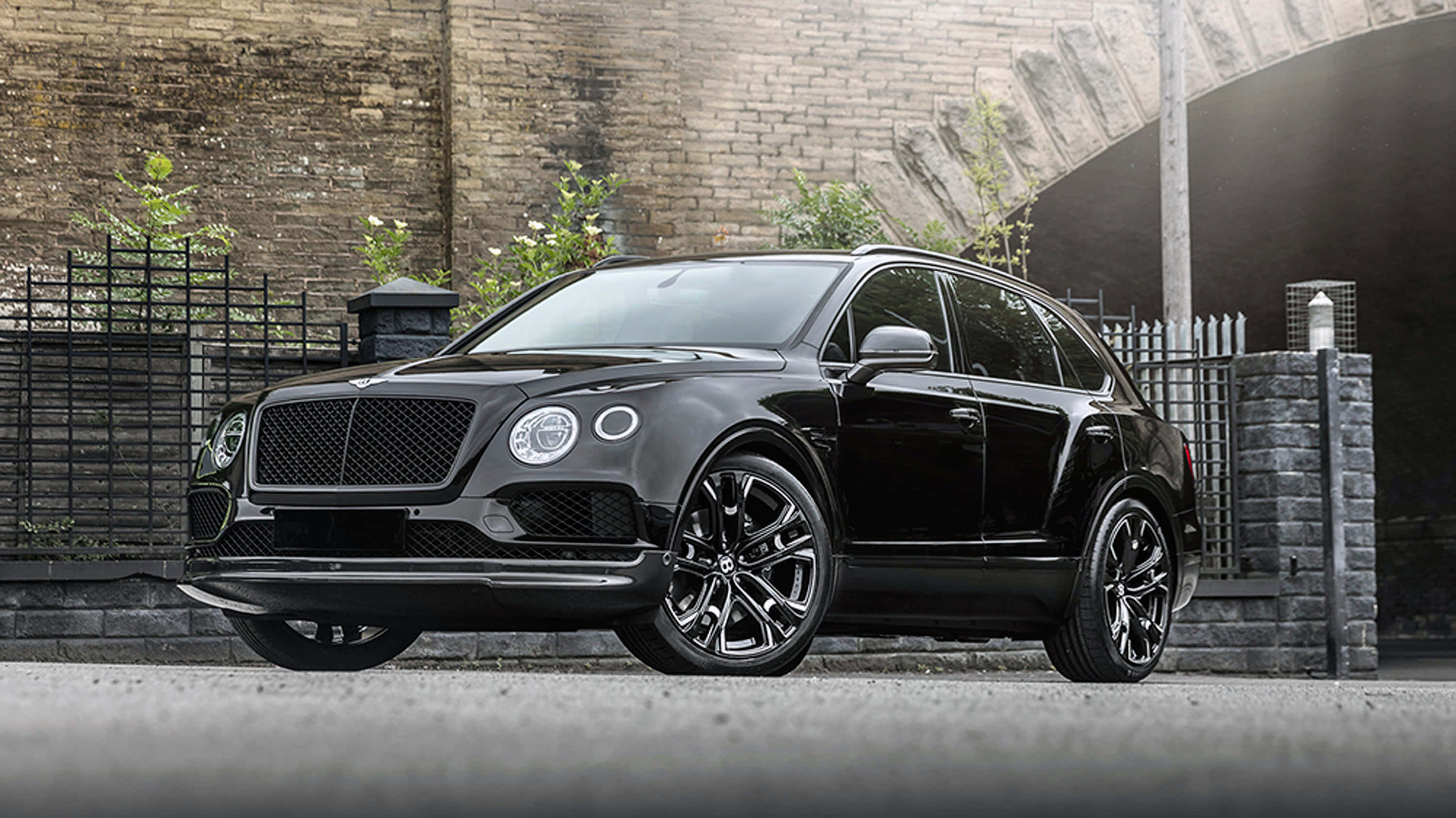Bentley Bentayga 桌布 壁紙 — 一輛時尚的黑色賓利Bentayga SUV停在風化的磚牆和金屬圍欄前。