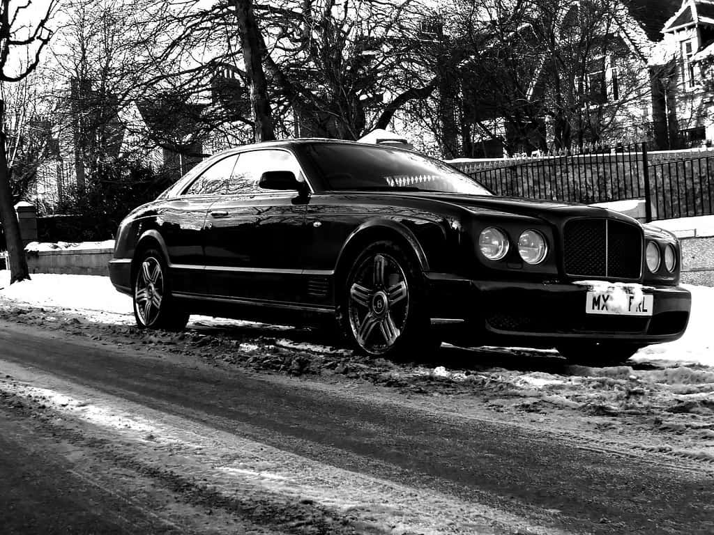 Bentley Brooklands 桌布 壁紙 — 一輛時尚的黑色賓利房車停在積雪街道上，背景有光禿的樹木和圍欄。