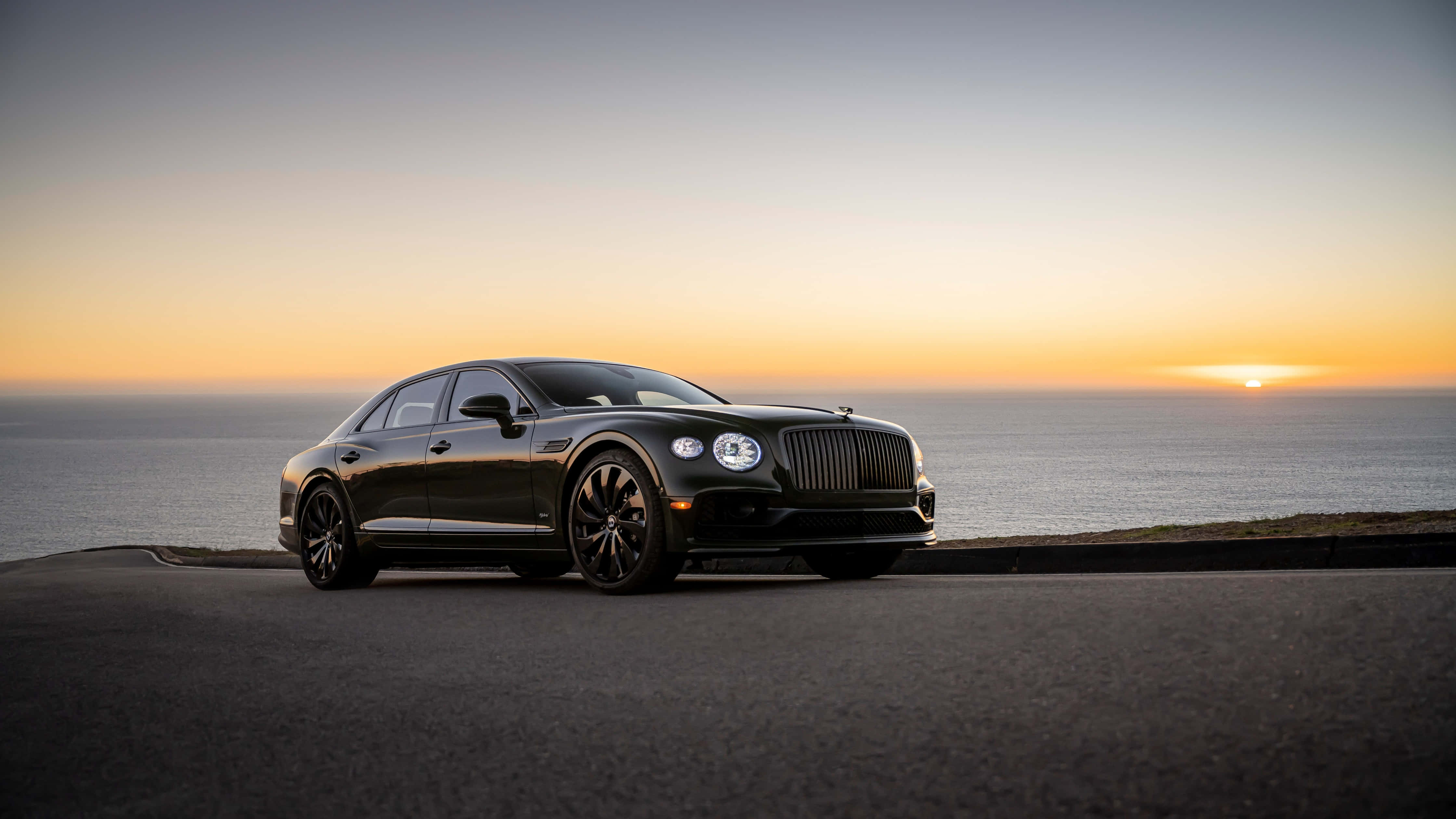 Bentley Flying Spur 桌布 壁紙 — 一輛時尚的黑色賓利房車停泊在日落時分的海岸公路上，海洋地平線閃耀著橙色光芒。