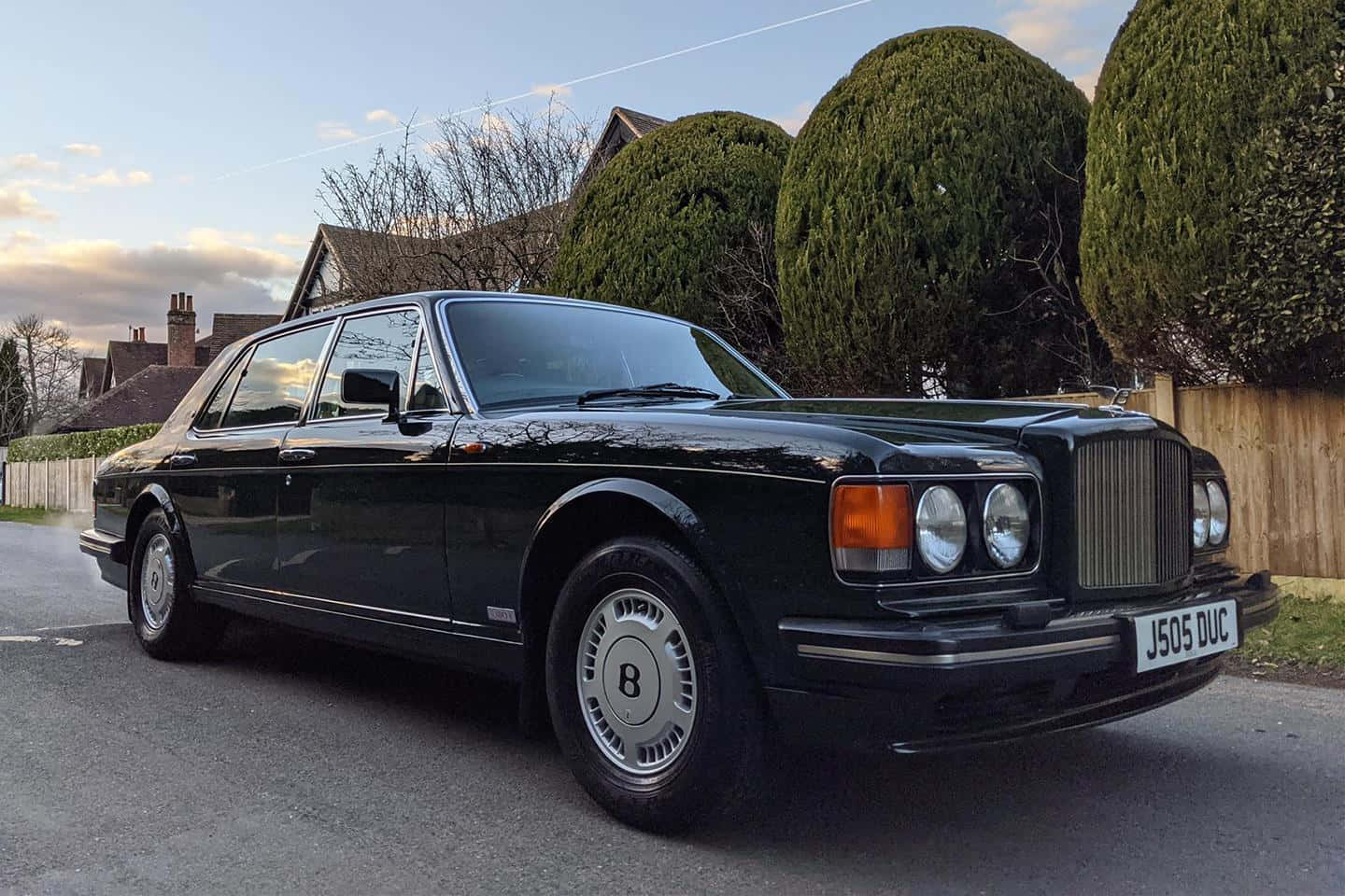 Bentley Turbo R 桌布 壁紙 — 一輛時尚的黑色賓利房車在黃昏時停泊在寧靜的住宅街道上，背景是修剪整齊的樹籬和房屋。