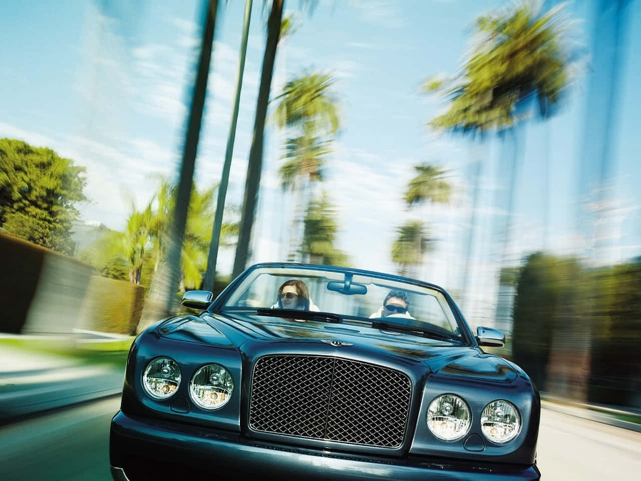 Bentley Azure 桌布 壁紙 — 一輛時尚的黑色賓利敞篷跑車在棕櫚樹成蔭的道路上行駛，車上兩人戴著太陽眼鏡，享受動態模糊的效果。