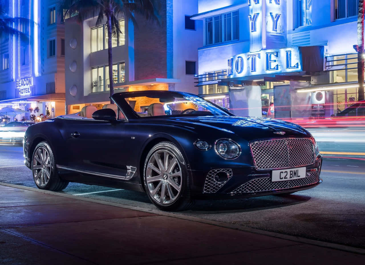 Bentley Continental Gt Convertible 桌布 壁紙 — 一輛時尚的黑色賓利敞篷車在夜晚停放在霓虹燈裝飾的Art Deco飯店前，周圍有棕櫚樹和模糊的城市燈光。