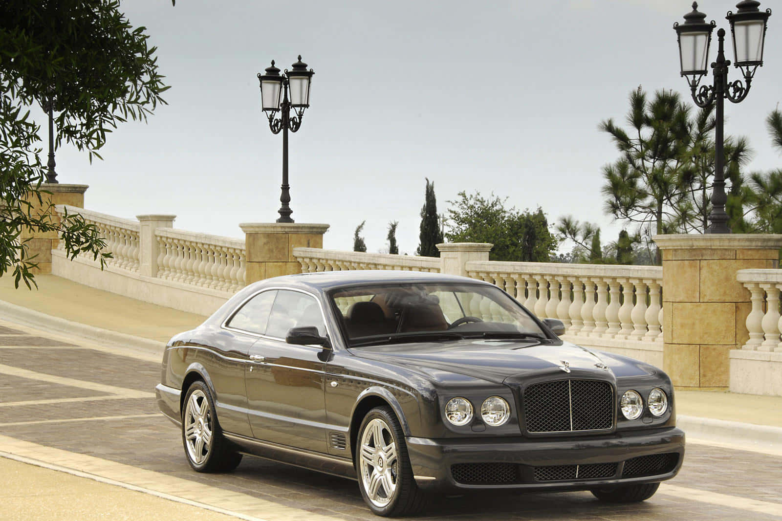 Bentley Brooklands 桌布 壁紙 — 一輛時尚的黑色賓利轎跑車在陽光明媚的石橋上滑行，兩旁是華麗的路燈和茂密的綠植。