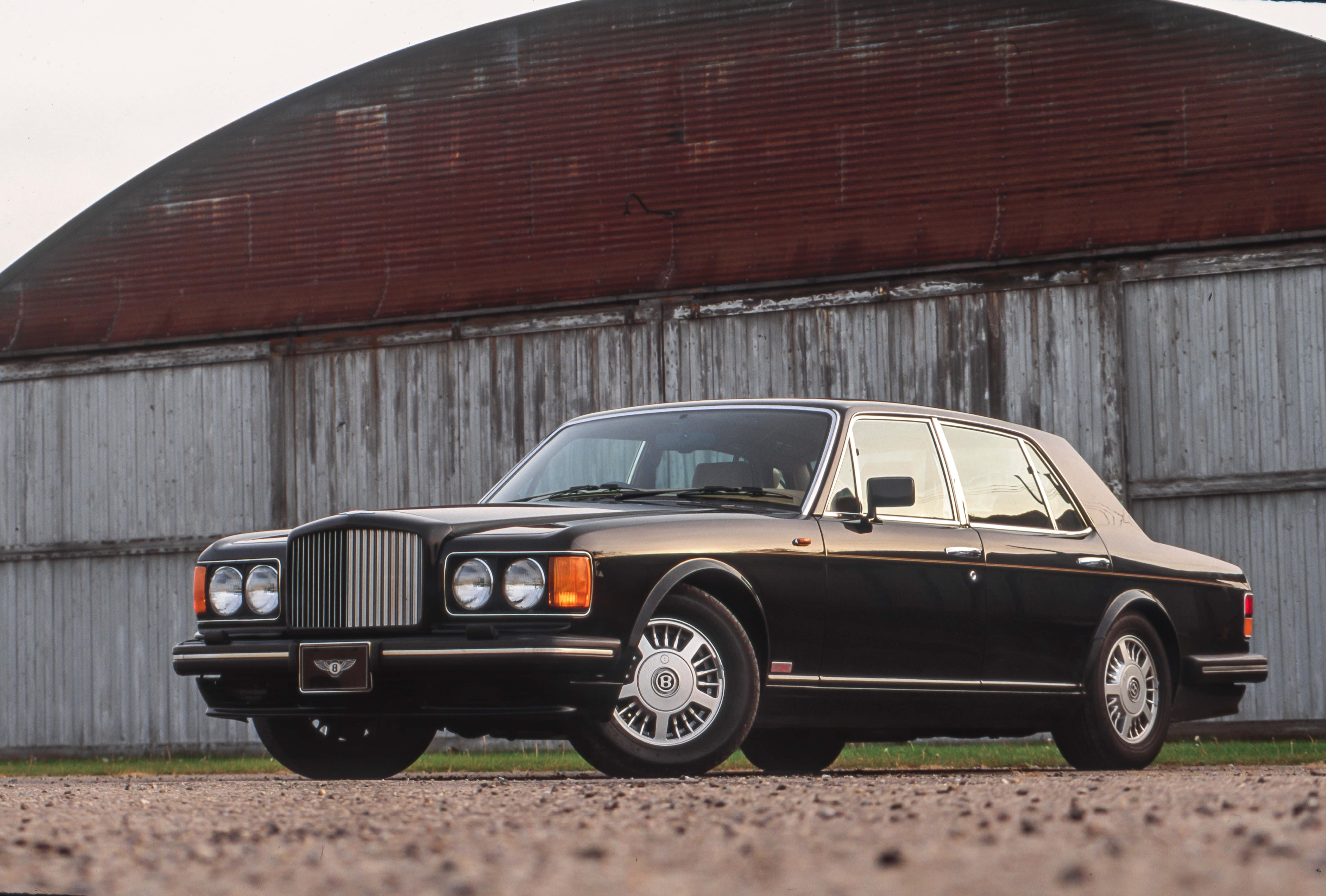 Bentley Turbo R 桌布 壁紙 — 一輛時尚的黑色賓利轎車停在碎石路上，前面是一座經風化的紅灰色穀倉，在柔和日光下。
