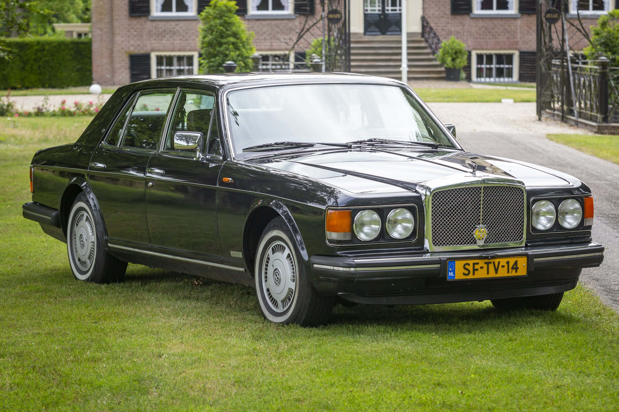 Bentley Eight 桌布 壁紙 — 一輛時尚的黑色賓利轎車停放在生氣勃勃的綠色草地上，前面是一座莊嚴的磚砌豪宅，配有修剪整齊的樹籬。