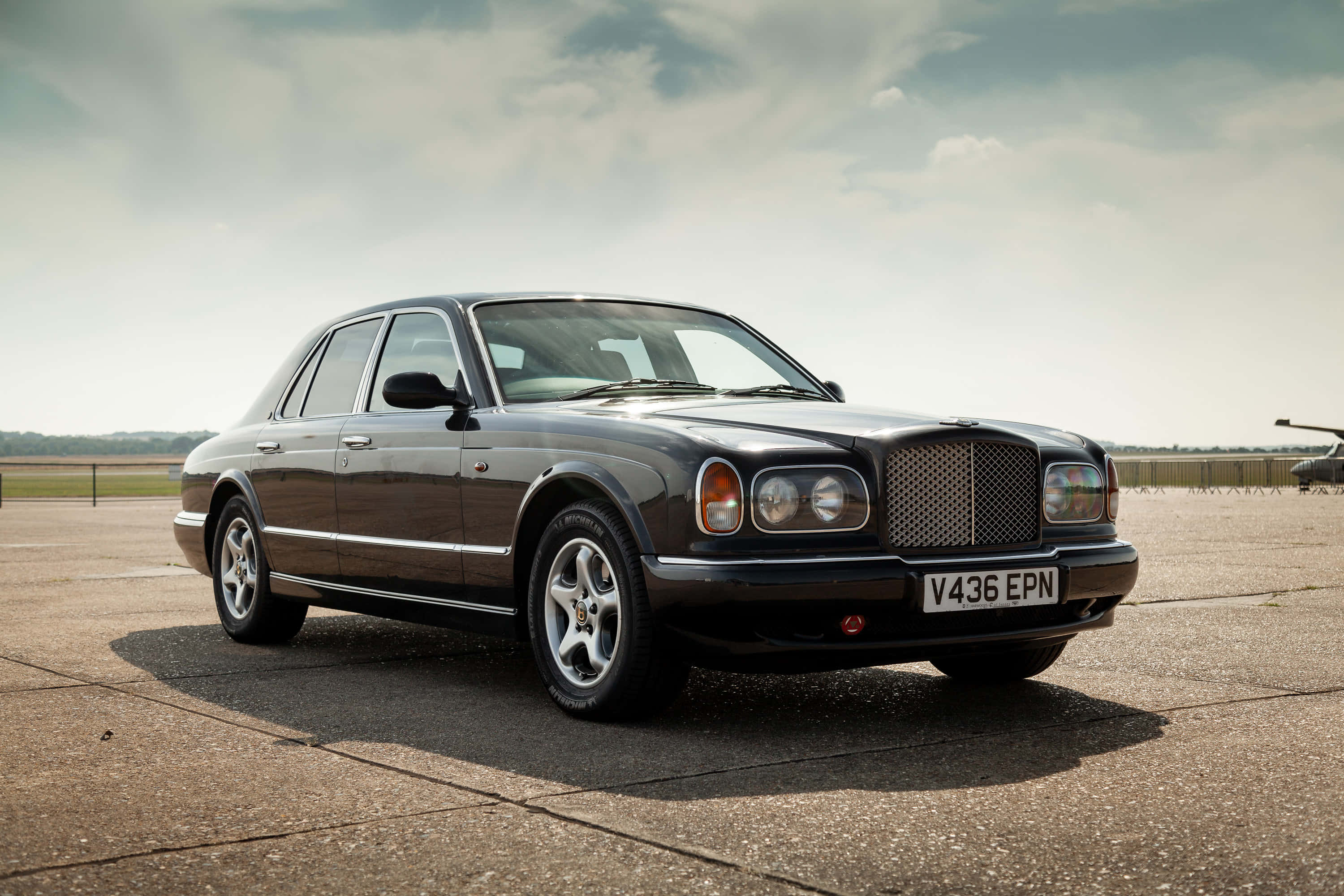 Bentley Arnage 桌布 壁紙 — 一輛時尚的黑色賓利轎車停放在鋪砌的機場上，天空多雲，遠處有樹木。