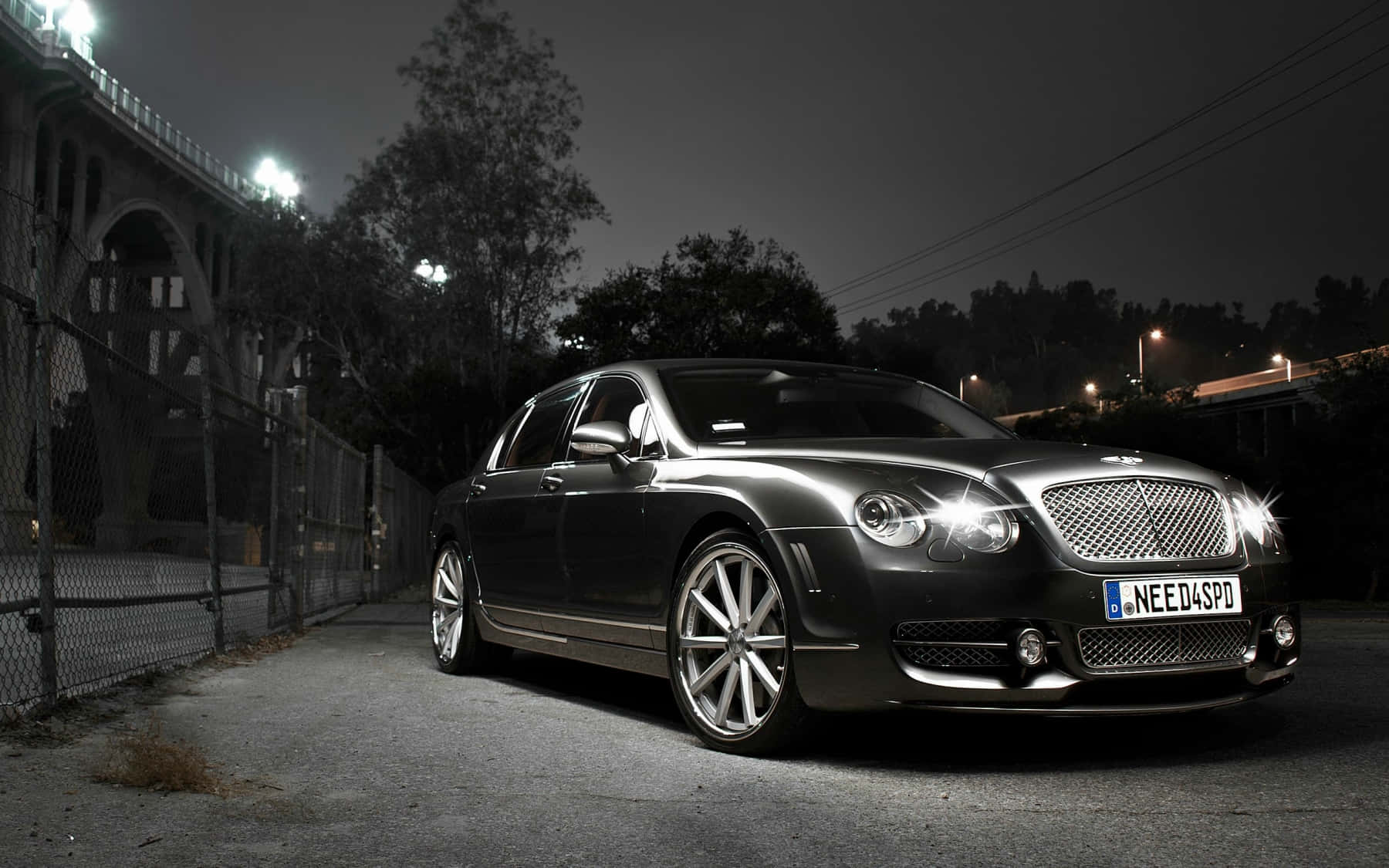 Bentley Flying Spur 桌布 壁紙 — 一輛時尚的黑色賓利轎車在人造燈光下閃閃發光，夜間停泊在鐵絲網圍欄和橋樑旁邊。