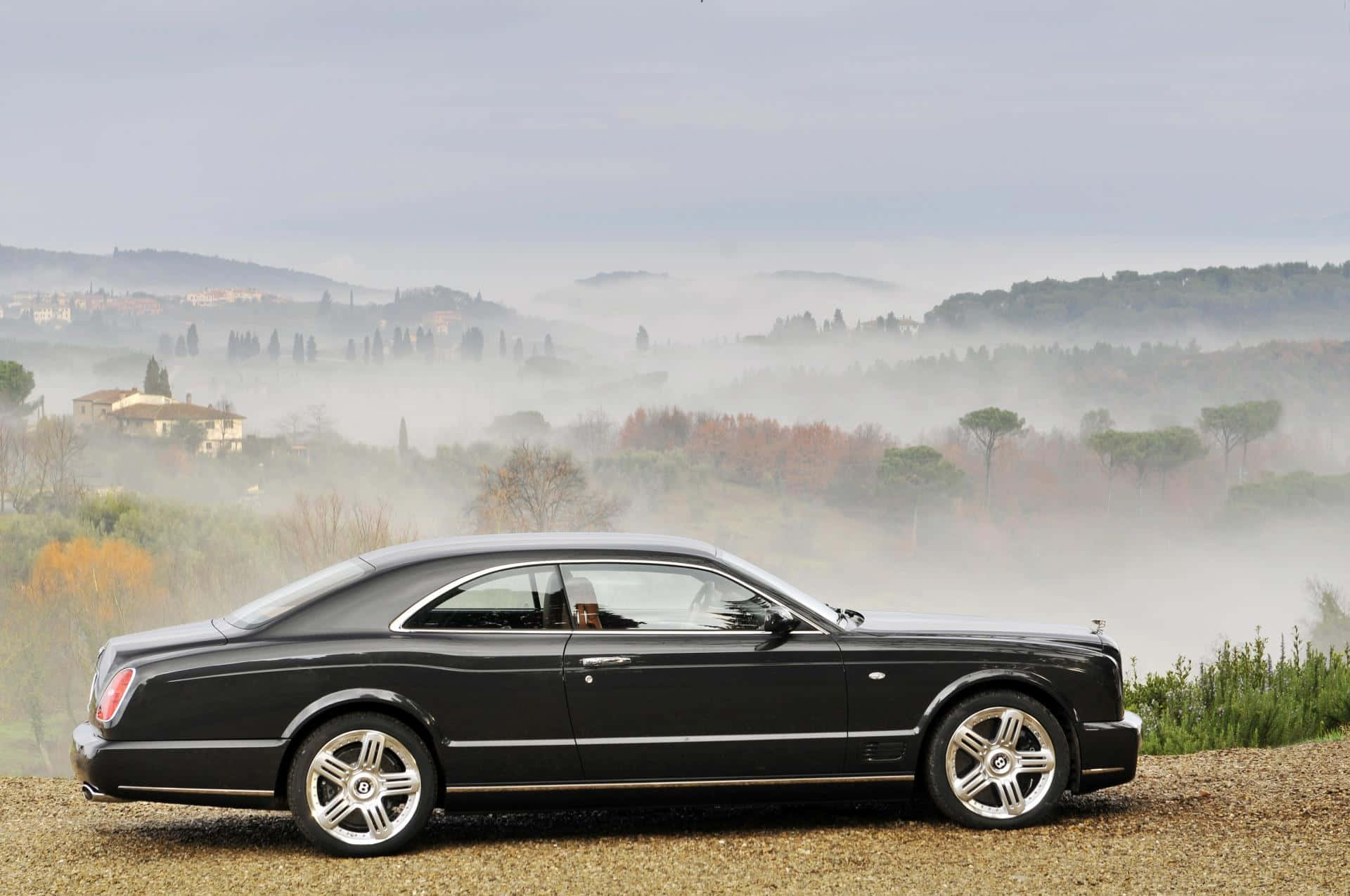 Bentley Brooklands 桌布 壁紙 — 一輛時尚的黑色賓利雙門跑車停泊在碎石觀景台上，薄霧籠罩著起伏的托斯卡納鄉村，點綴著樹木和遠處的別墅。