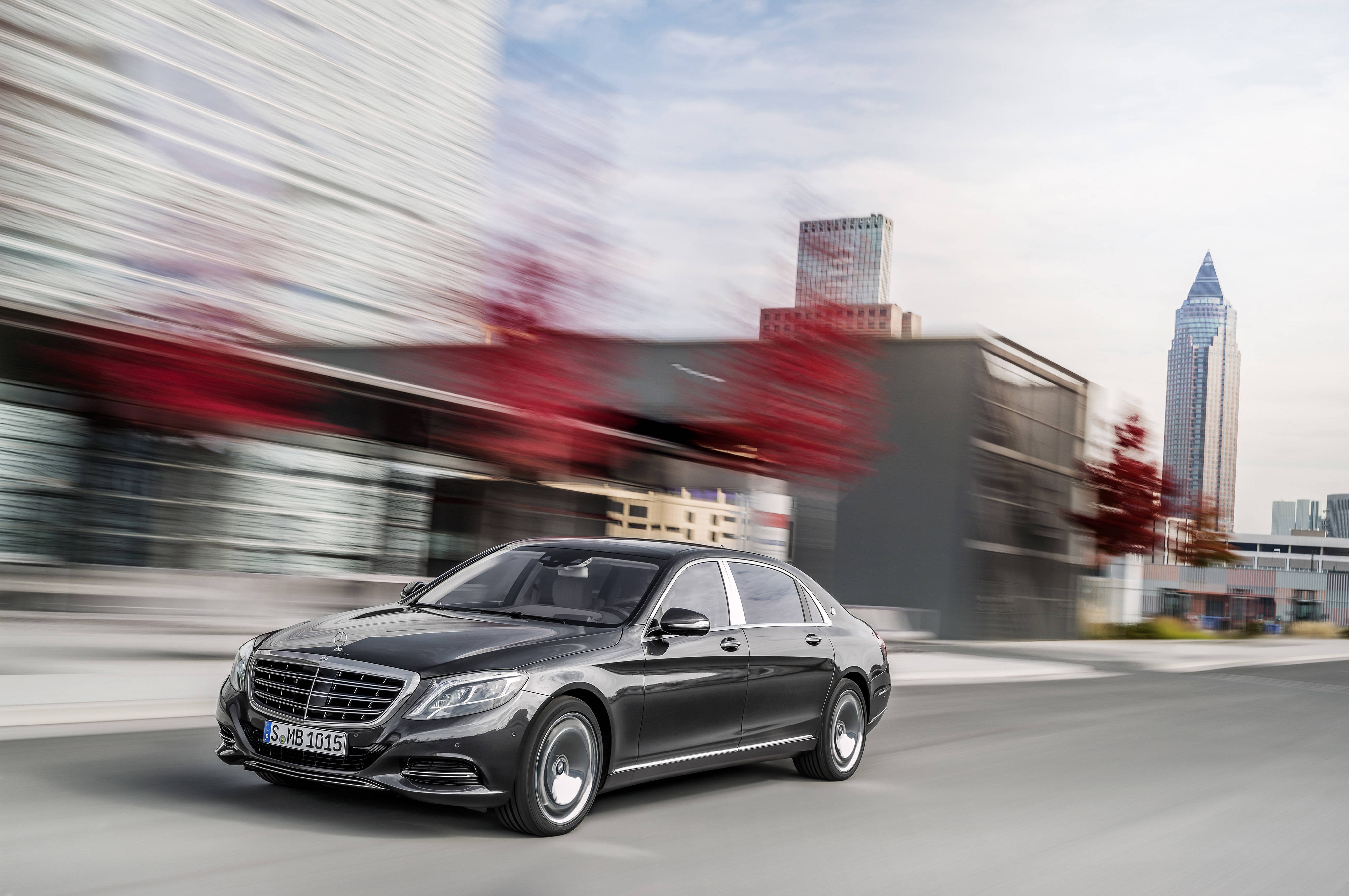 benz 4K 桌布 壁紙 — 一輛時尚的黑色賓士S-Class轎車在現代城市景觀中疾馳，建築物和紅樹呈現動態模糊效果。