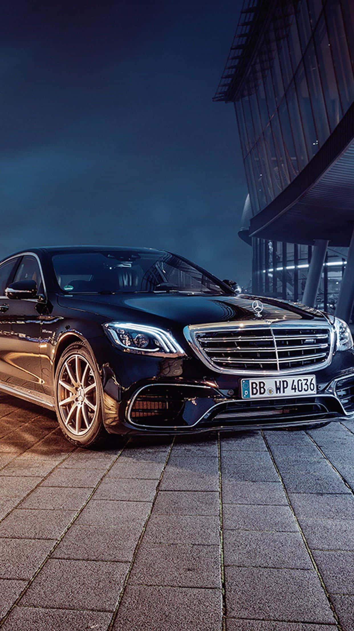 Mercedes 桌布 壁紙 — 一輛時尚的黑色賓士S-Class轎車在夜燈下閃耀，旁邊是現代玻璃建築，位於石鋪路面上。