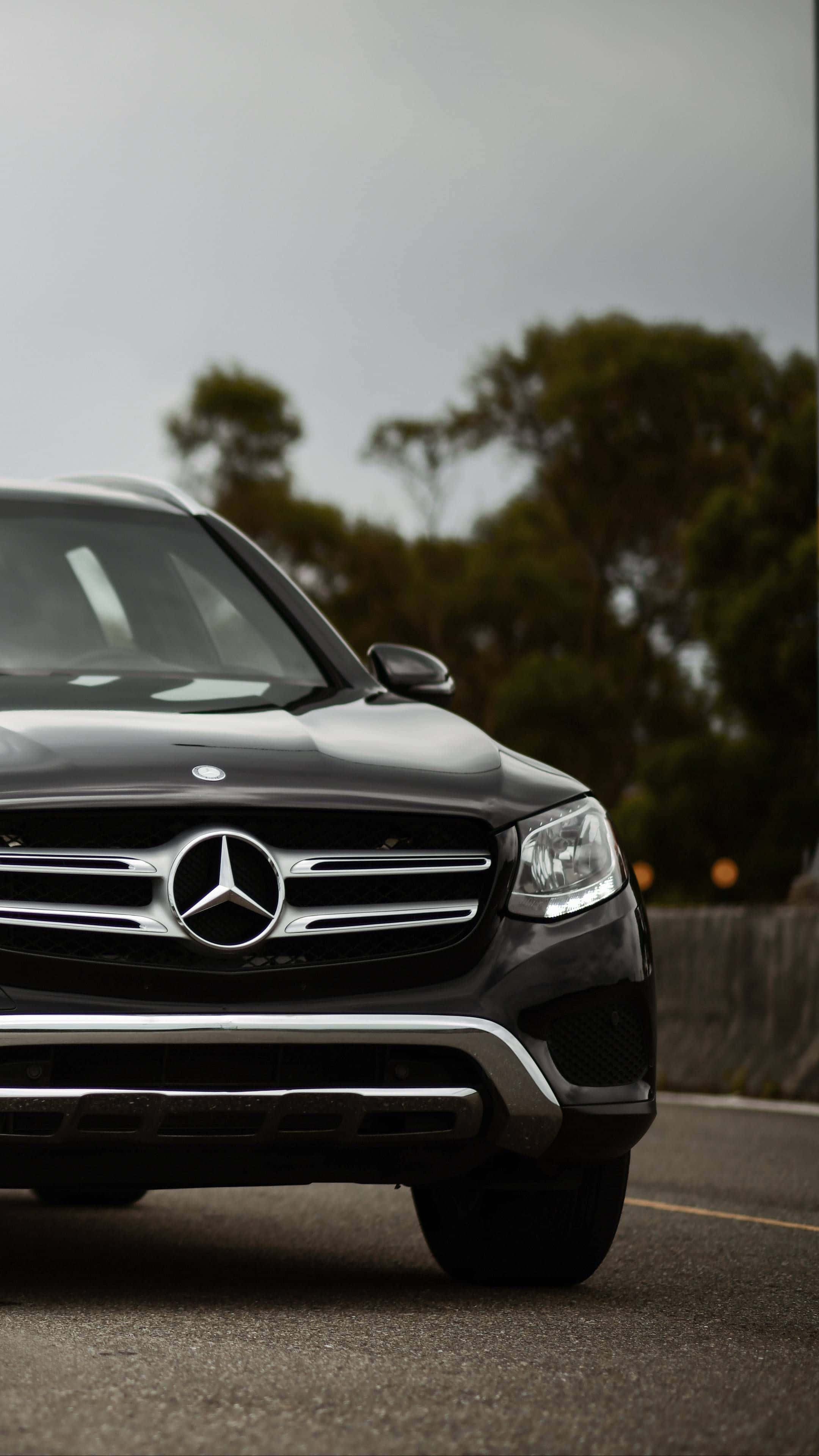 Mercedes 桌布 壁紙 — 一輛時尚的黑色賓士SUV停在柏油路上，背景是模糊的綠樹，天空多雲。