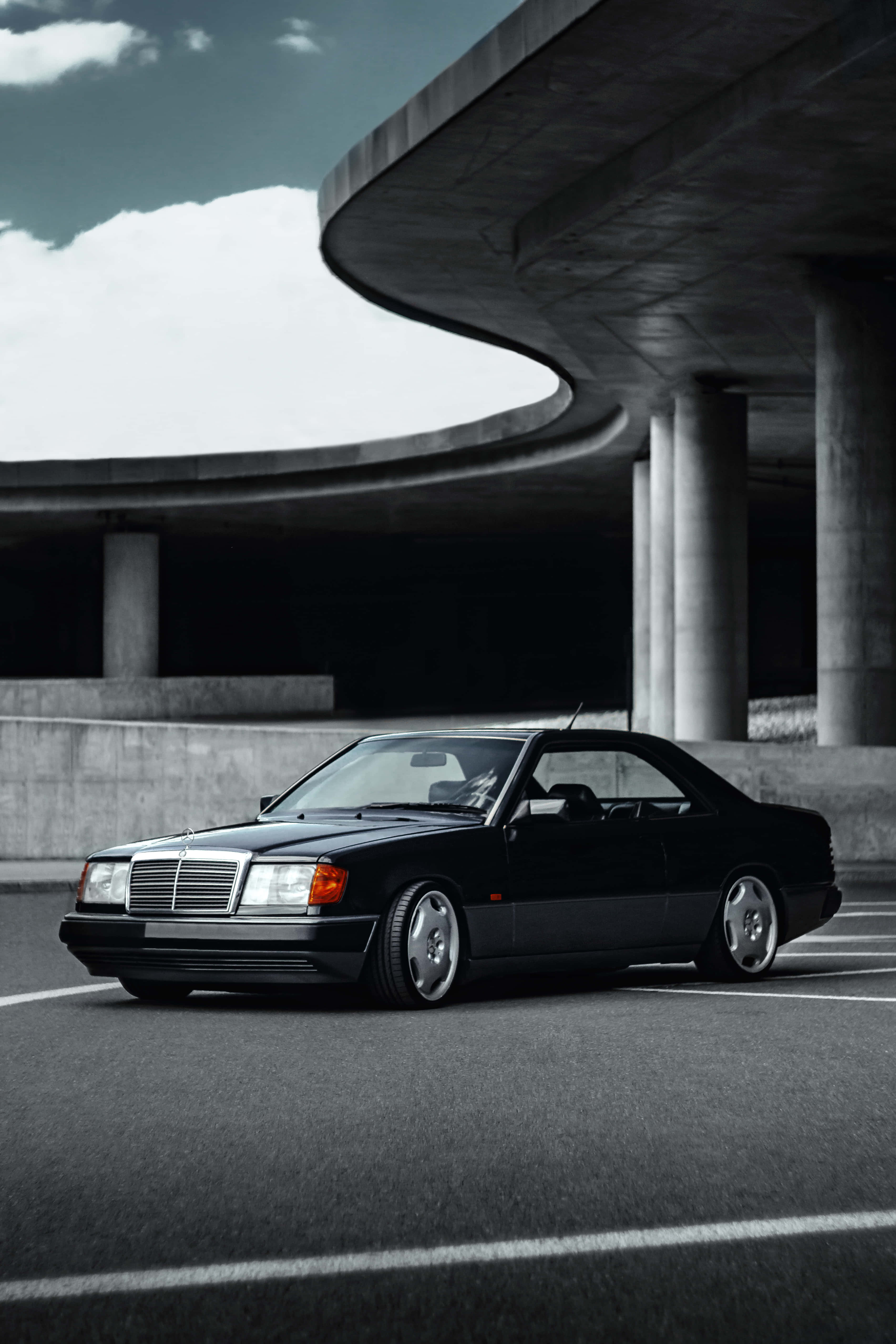 Classic Mercedes 桌布 壁紙 — 一輛時尚的黑色賓士轎跑車停放在彎曲的混凝土天橋下，天空多雲，氣氛戲劇化。