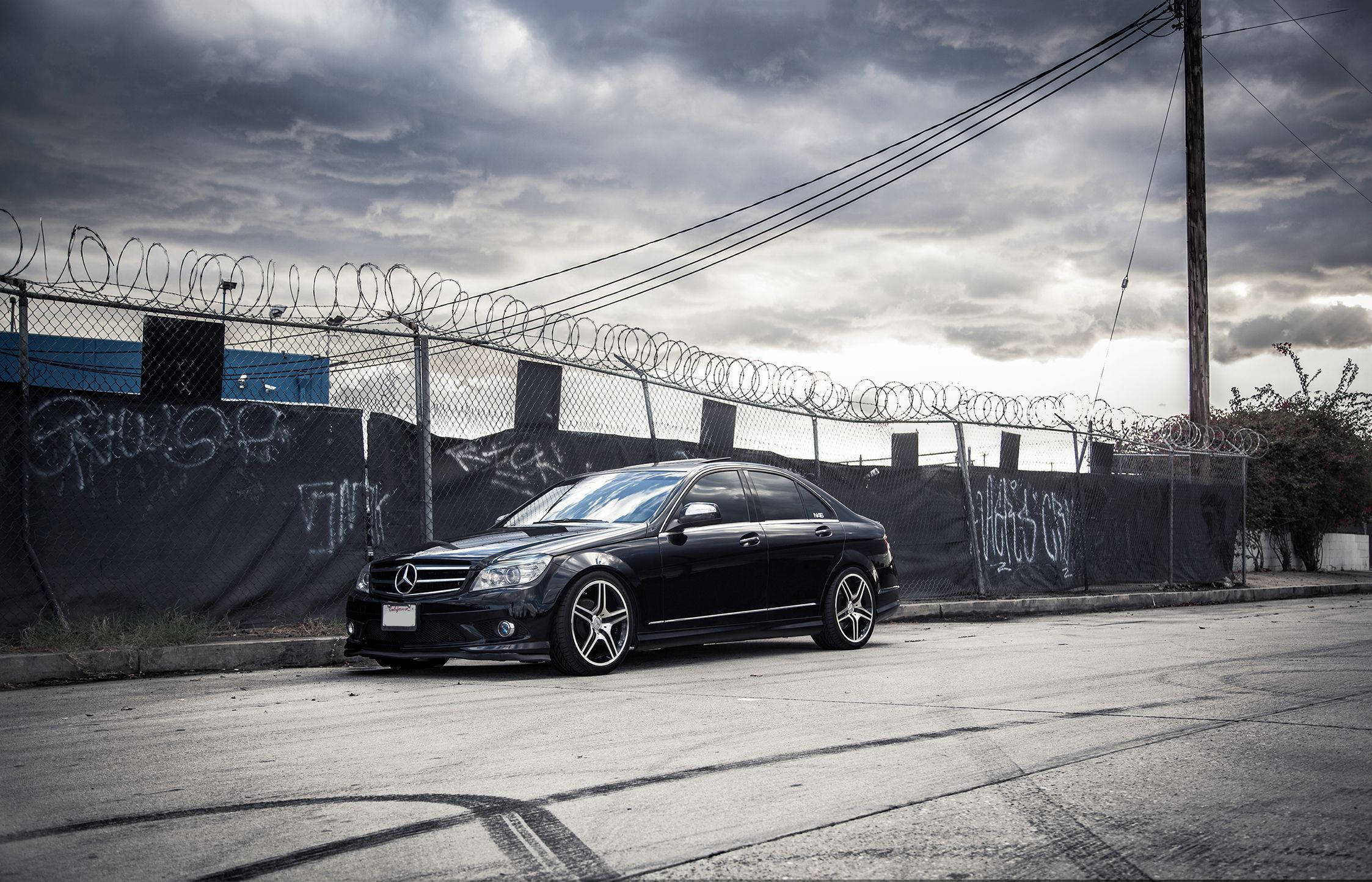 Mercedes Benz C300 桌布 壁紙 — 一輛時尚的黑色賓士轎車停在裂開的柏油路上，旁邊是佈滿塗鴉的圍欄，頂部有鐵絲網，天空風雨欲來。