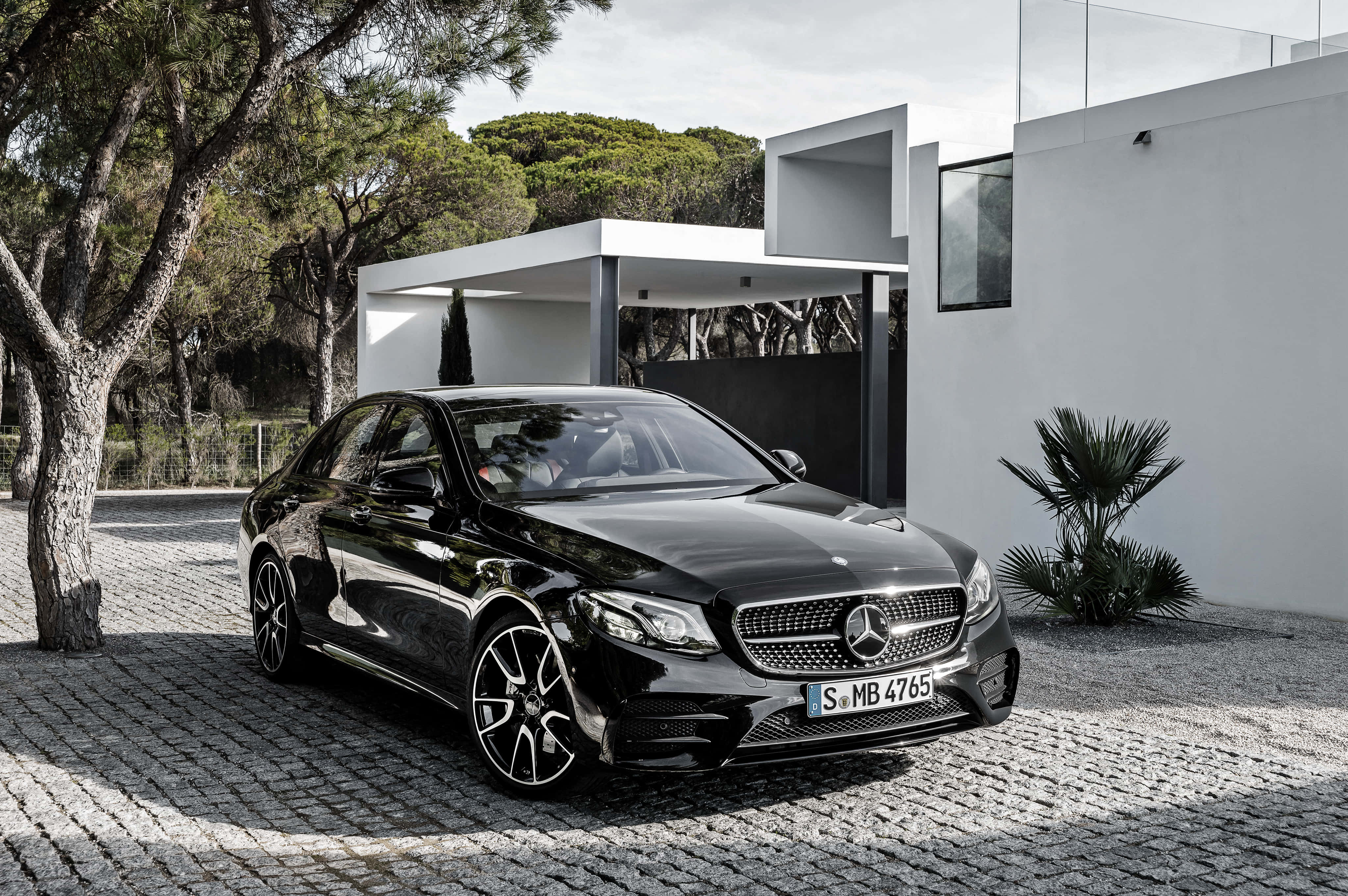 Mercedes 桌布 壁紙 — 一輛時尚的黑色賓士轎車在日光下閃耀，停泊在現代白色房屋旁邊的鵝卵石車道上，背景有樹木。