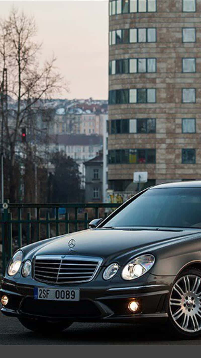 Black Car 桌布 壁紙 — 一輛時尚的黑色賓士轎車在柔和暮光下閃閃發光，旁邊是一座現代磚砌建築，背景有光禿禿的樹木。