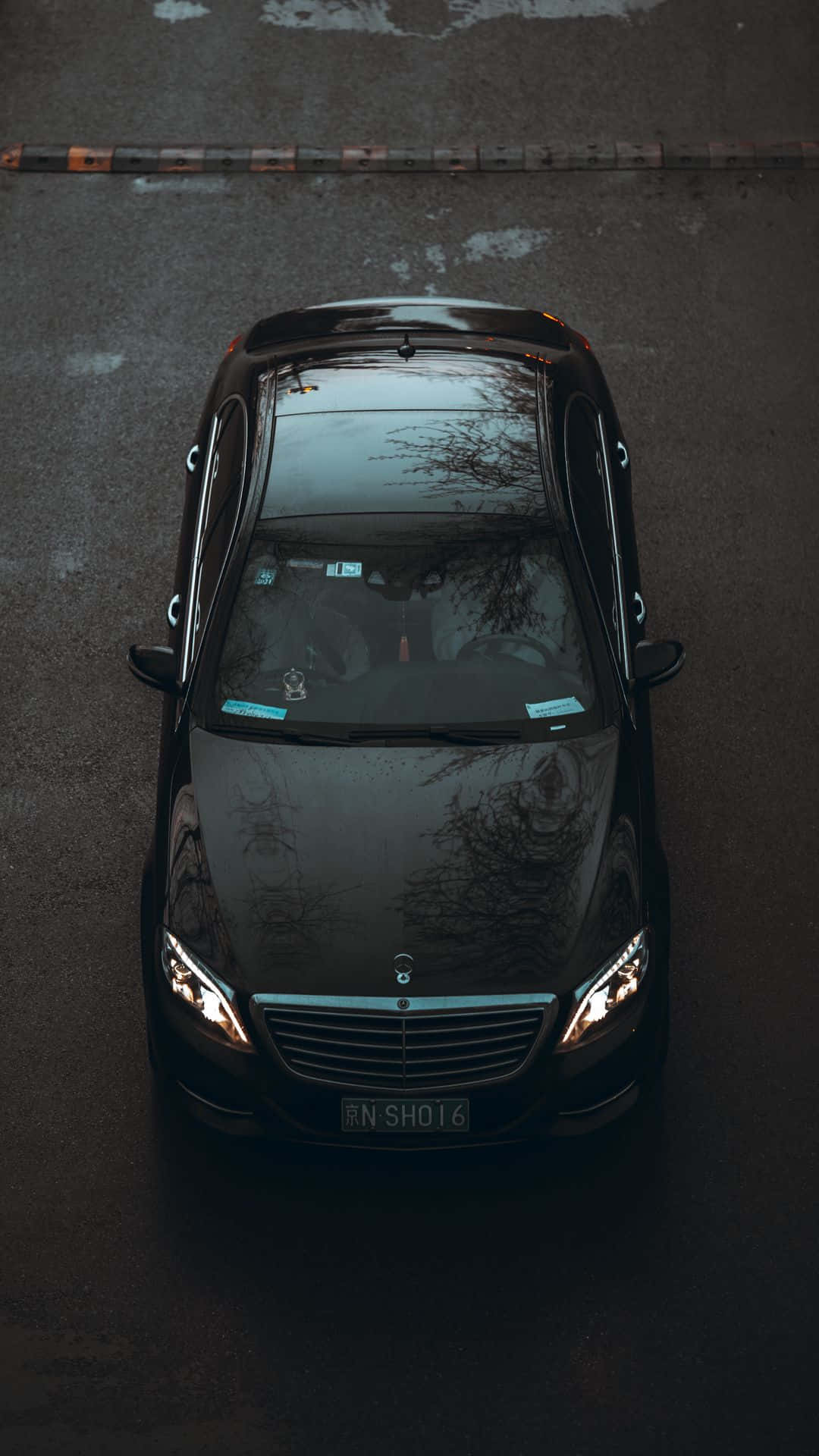 Mercedes 桌布 壁紙 — 一輛時尚的黑色賓士轎車從上方俯瞰，停在濕漉漉的柏油街道上，引擎蓋上反射著樹木的倒影。