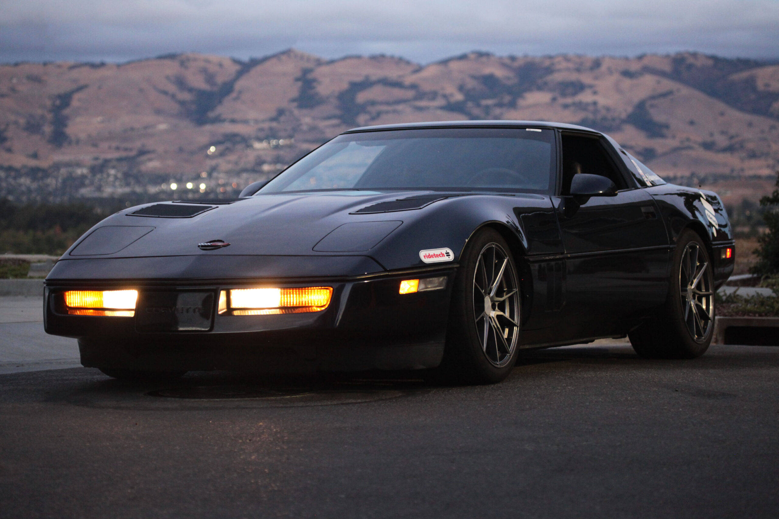 corvette 桌布 壁紙 — 一輛時尚的黑色跑車，帶有發光車頭燈，停在瀝青路面上，背景是黃昏時分起伏的山丘。