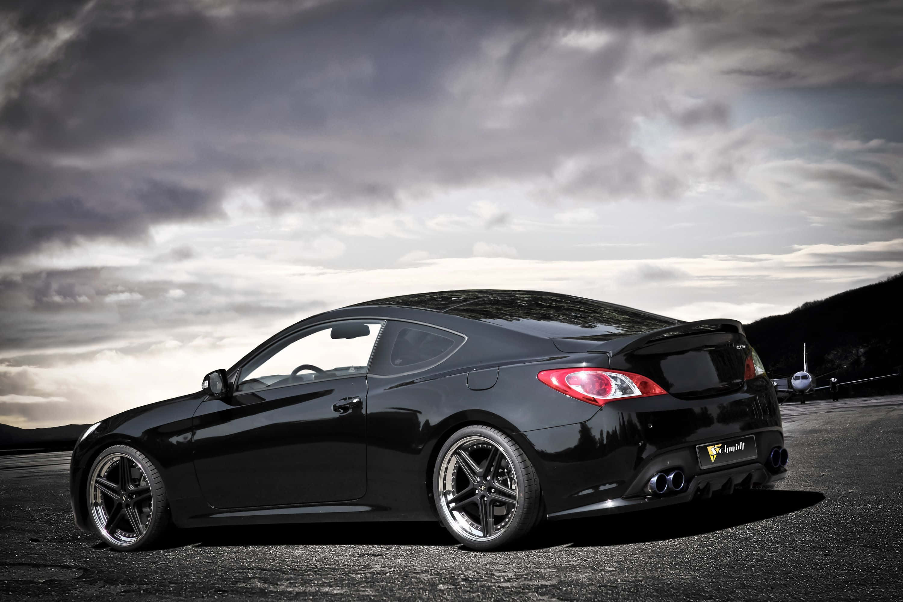 Hyundai Genesis 桌布 壁紙 — 一輛時尚的黑色跑車，配備定制輪圈和雙排氣管，停在戲劇性多雲天空下的跑道上。