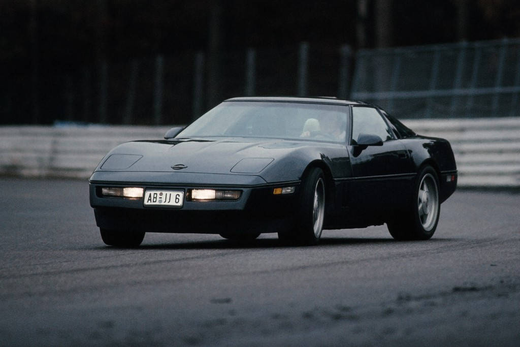 corvette 桌布 壁紙 — 一輛時尚的黑色跑車，配備發光的車頭燈，沿著一條濕滑的瀝青賽道疾馳，賽道兩旁設有安全護欄。