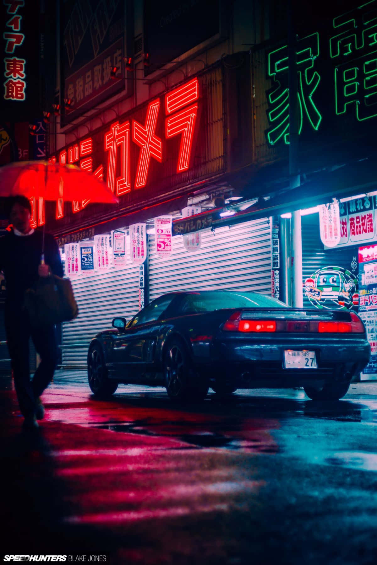 JDM iPhone 桌布 壁紙 — 一輛時尚的黑色跑車停在夜晚霓虹燈閃爍的東京雨街上，一個拿著紅色雨傘的人走過。