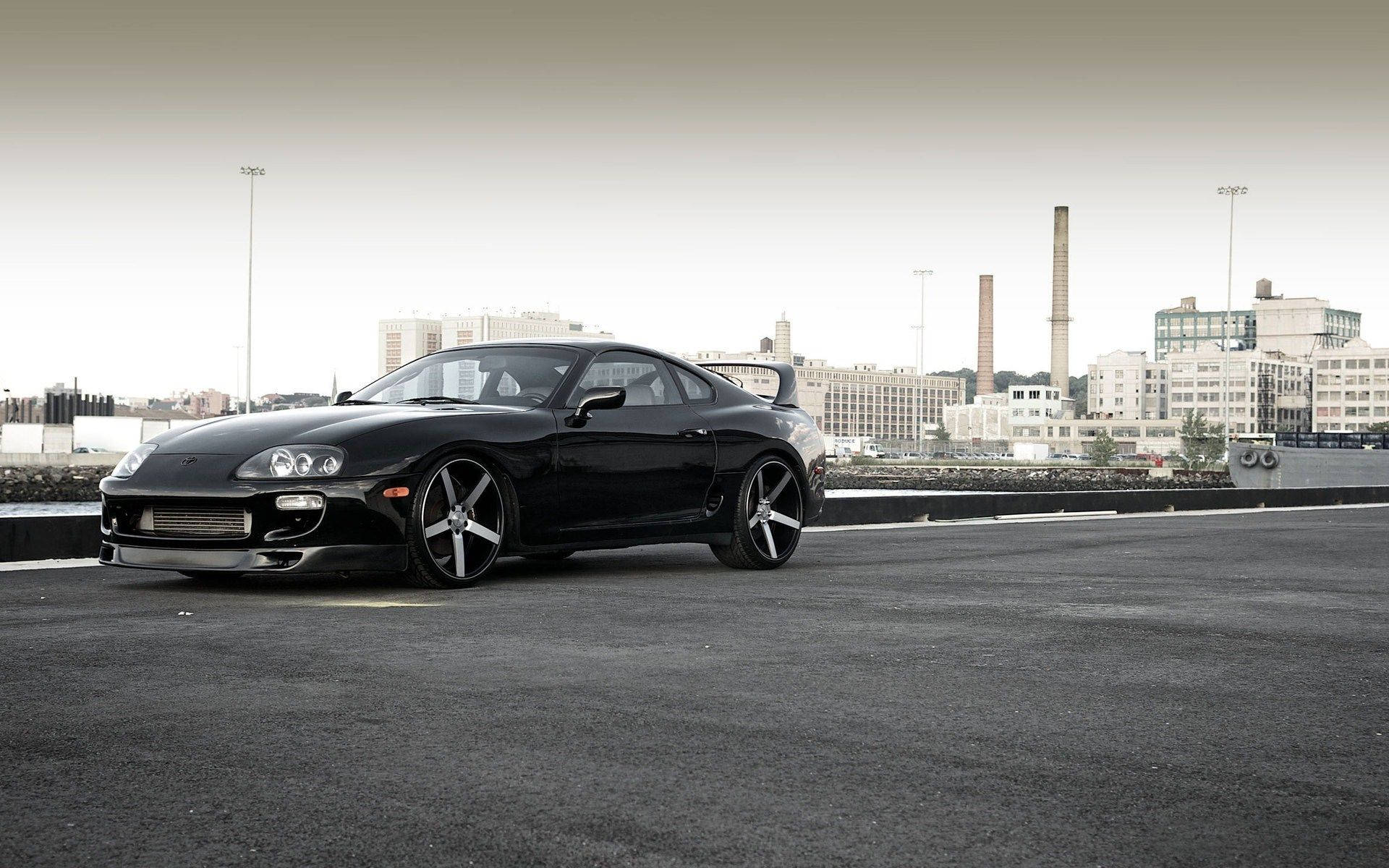 黑色跑車停在瀝青上，背後有城市和工業建築。 — Toyota Supra Mk4 壁紙