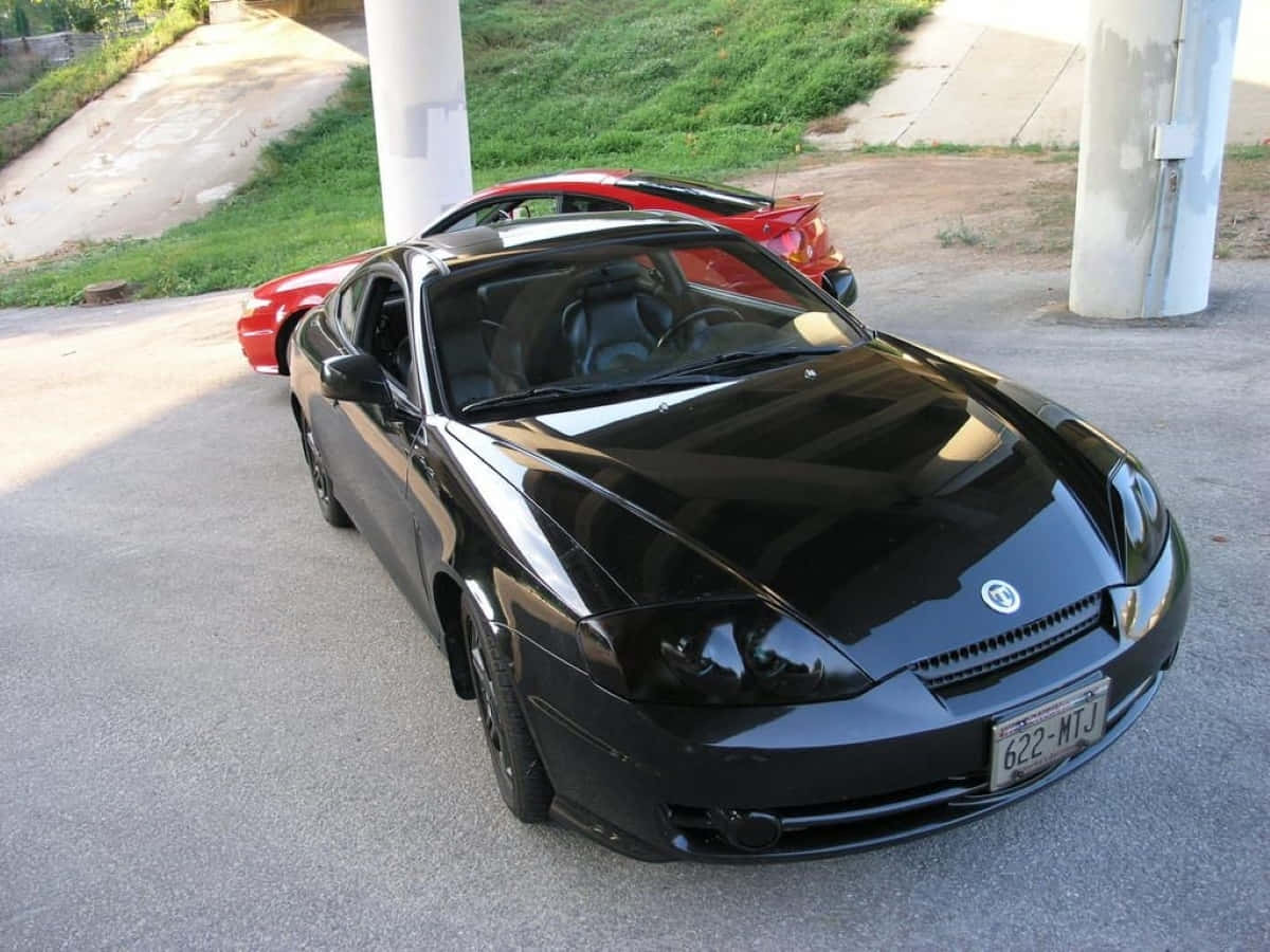 Hyundai Tiburon 桌布 壁紙 — 一輛時尚的黑色跑車停放在混凝土柱下，後面有一輛紅色汽車，在陽光明媚的日子裡。