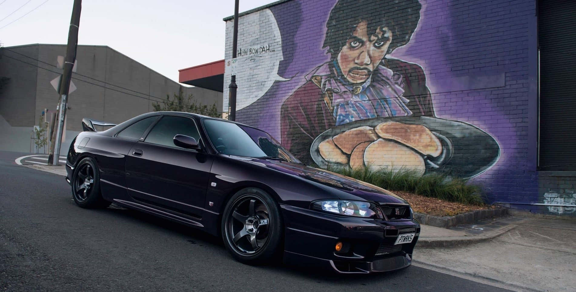 R33 GT-R 桌布 壁紙 — 一輛時尚的黑色跑車停放在街頭一幅Prince手持巨型麵包的鮮紫色壁畫旁邊。