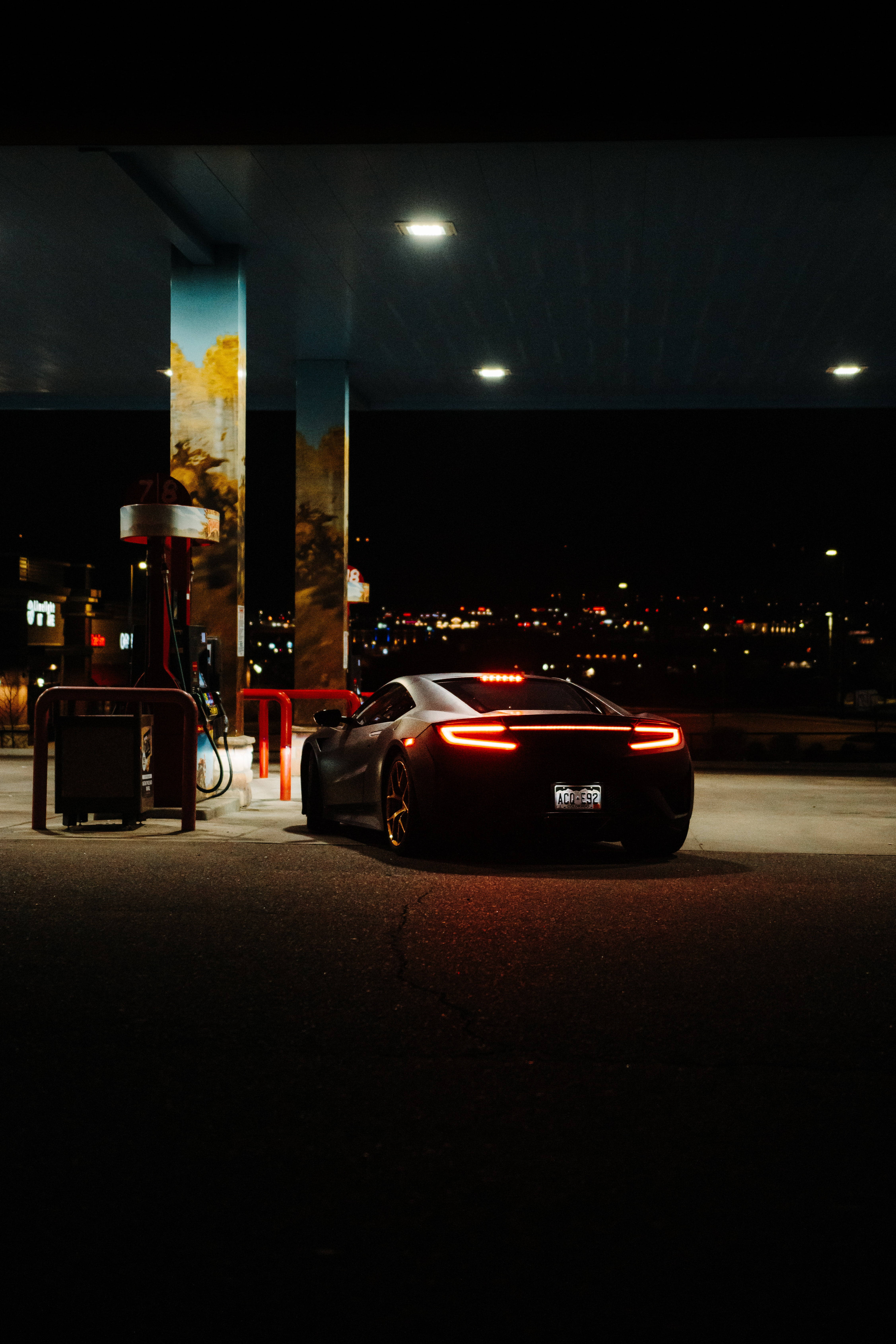 Gas Station 桌布 壁紙 — 一輛時尚的黑色跑車在夜空中閃爍著紅色尾燈，位於加油站，背景有遠處的城市燈光。