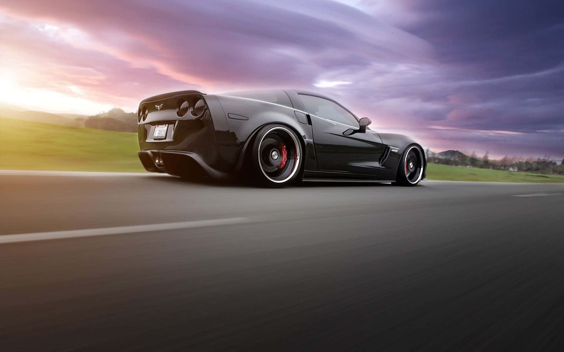 Chevrolet Corvette Z06 桌布 壁紙 — 一輛時尚的黑色跑車在戲劇性的紫色和橙色日落天空下沿著道路飛馳。