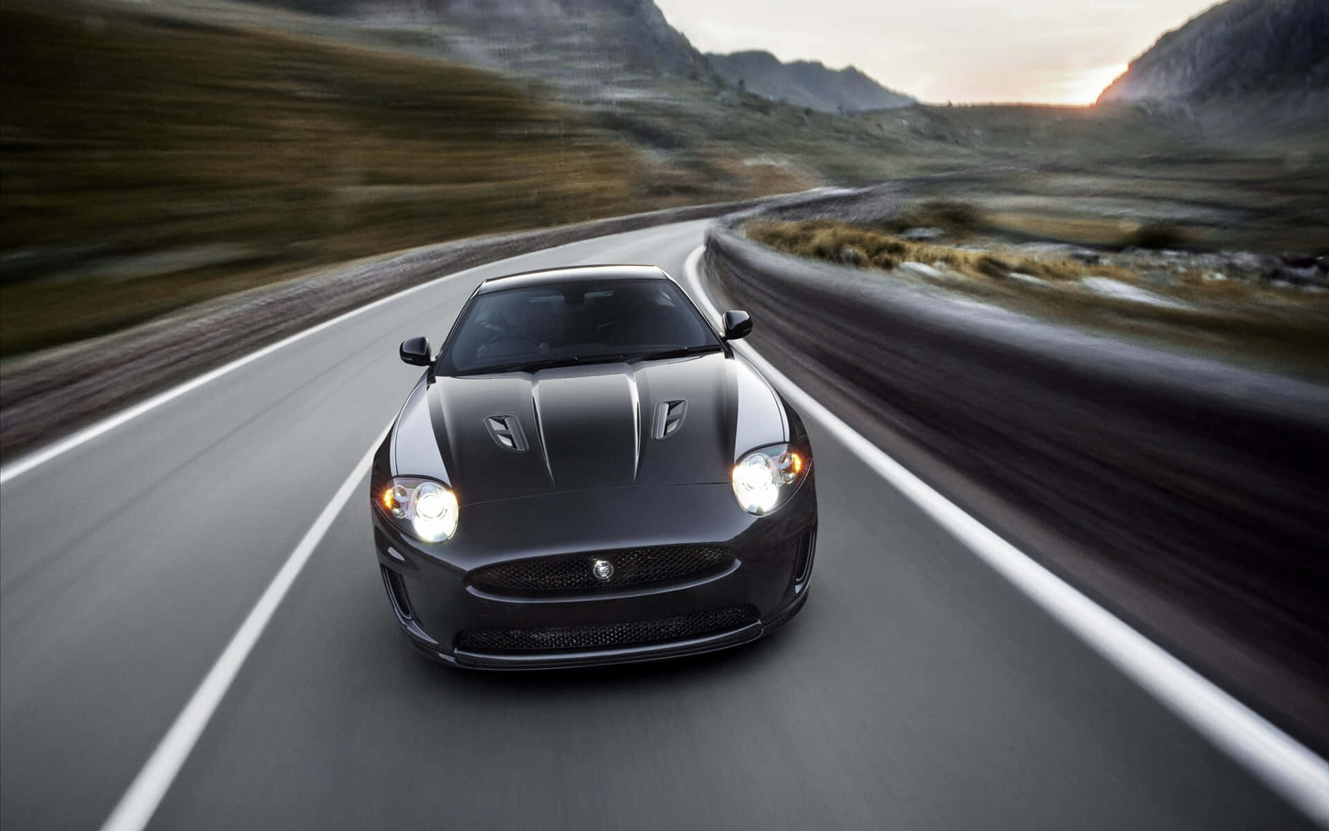 Jaguar Xkr 桌布 壁紙 — 一輛時尚的黑色跑車在日落時分沿著蜿蜒的山路疾馳，背景模糊。