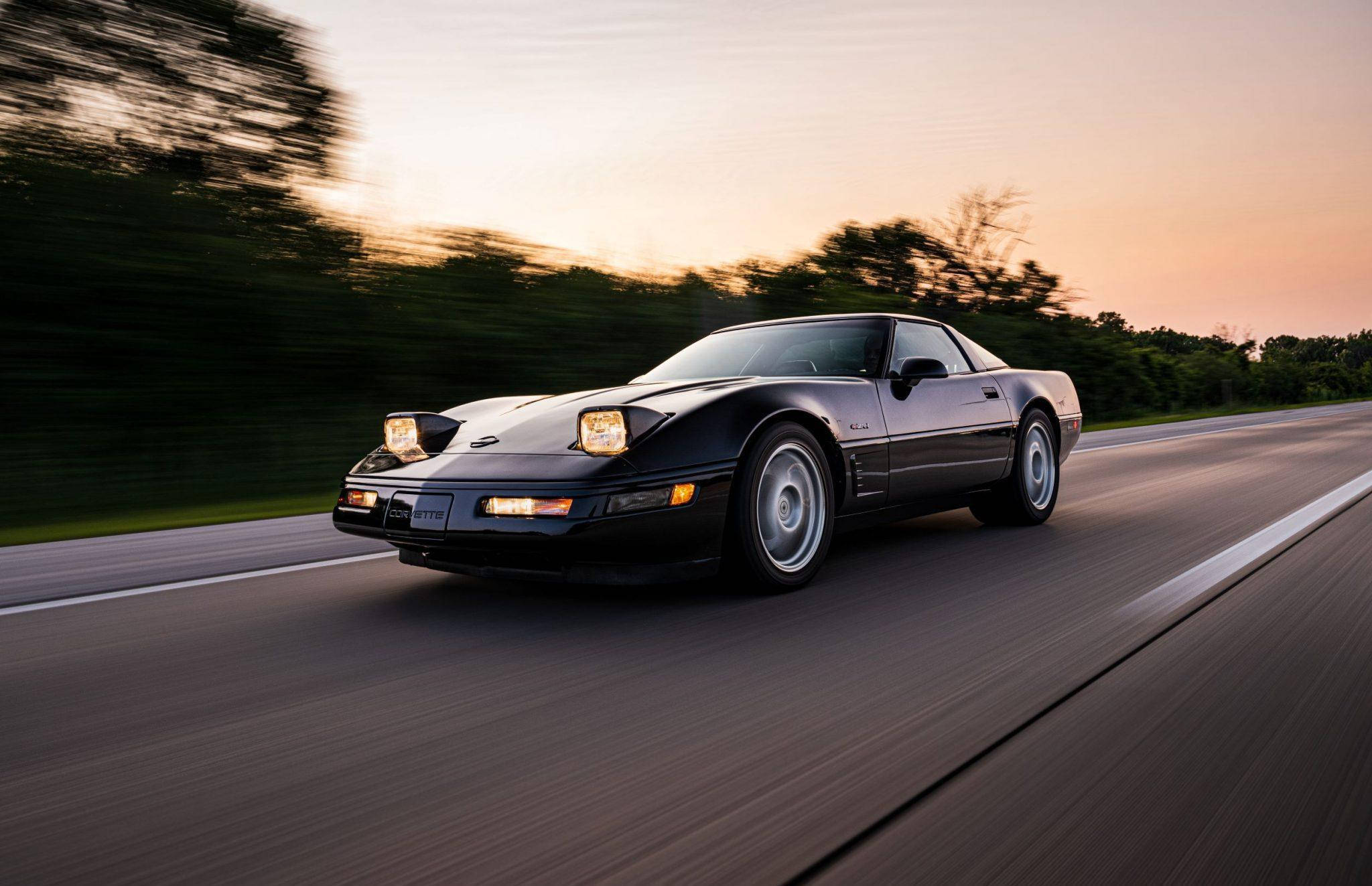 corvette 桌布 壁紙 — 一輛時尚的黑色跑車在日落時分的高速公路上疾馳，背景是模糊的樹木和溫暖的金色光芒。