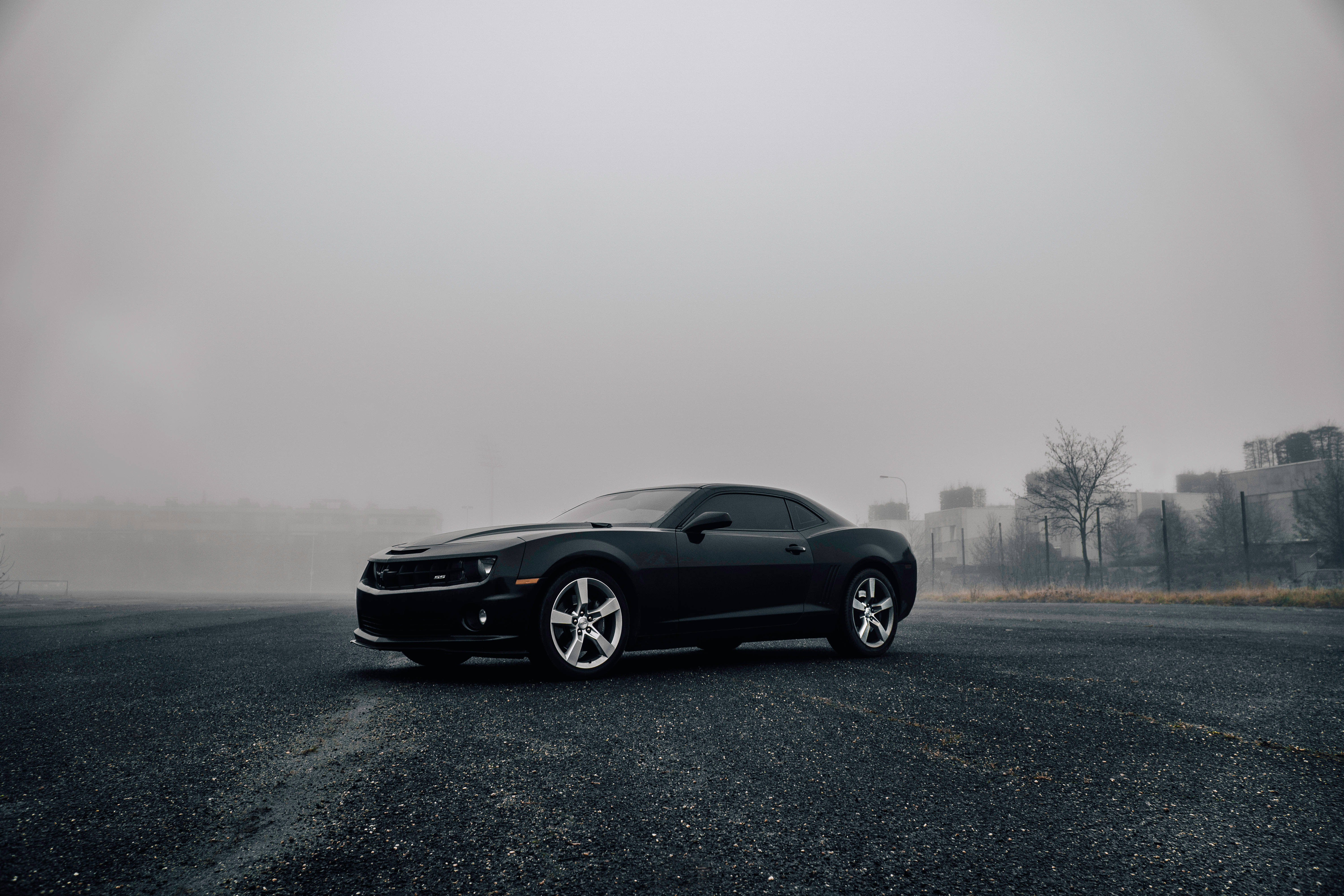 Chevrolet 桌布 壁紙 — 一輛時尚的黑色跑車孤獨地停在霧氣瀰漫的瀝青停車場上，灰色調柔和，遠處建築物在霧中幾乎看不見。