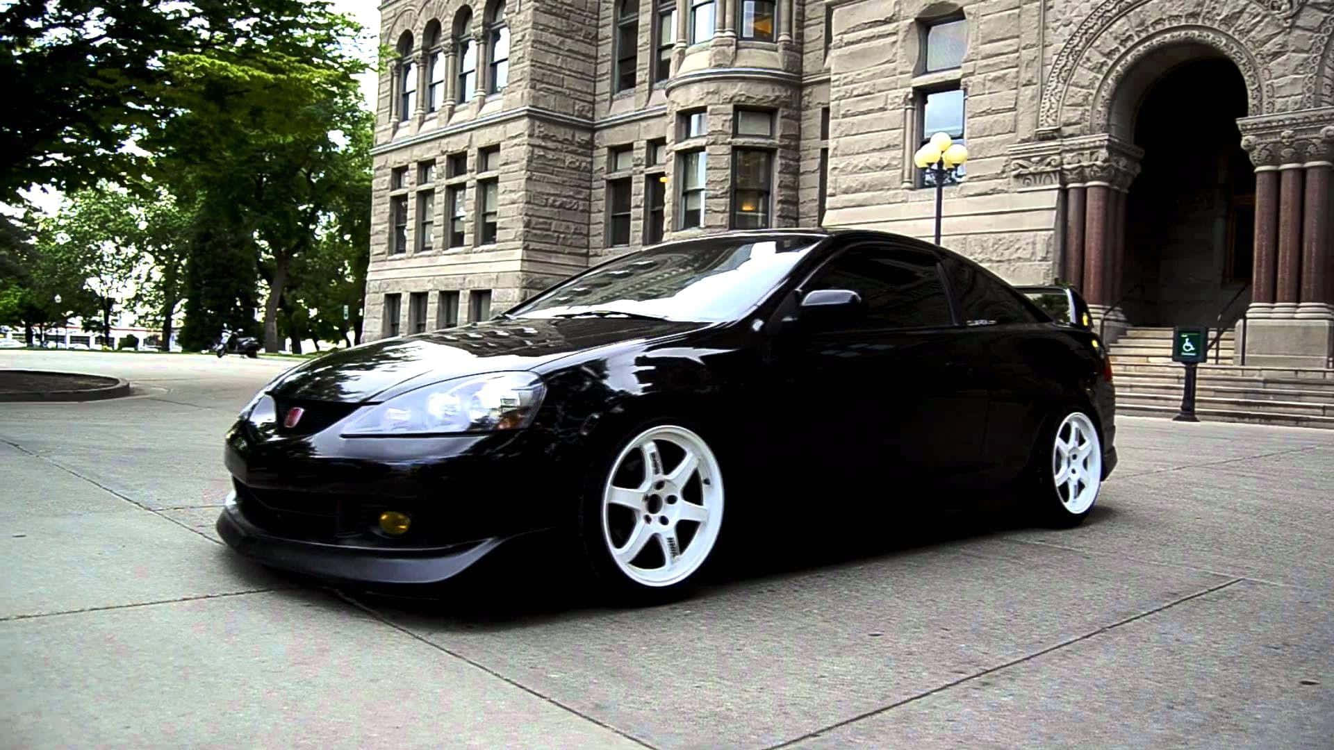 Rsx Type-s 桌布 壁紙 — 一輛時尚的黑色跑車搭配白色輪胎，停放在鋪砌的廣場上，前方是一座宏偉的石造建築，帶有拱形入口和樹木。