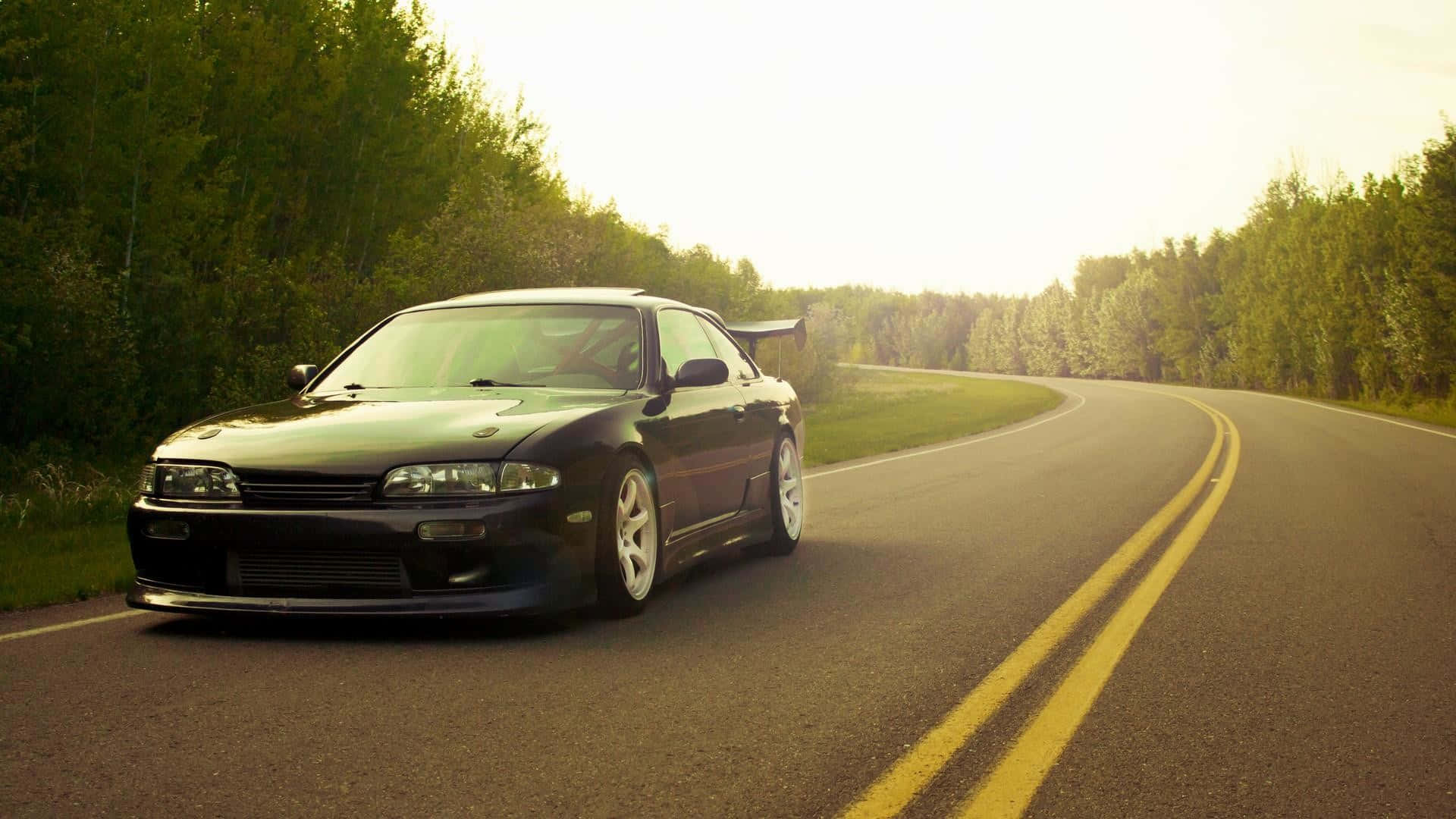 JDM 桌布 壁紙 — 一輛時尚的黑色跑車沿著蜿蜒的森林道路行駛，沐浴在金色的日落光芒中。