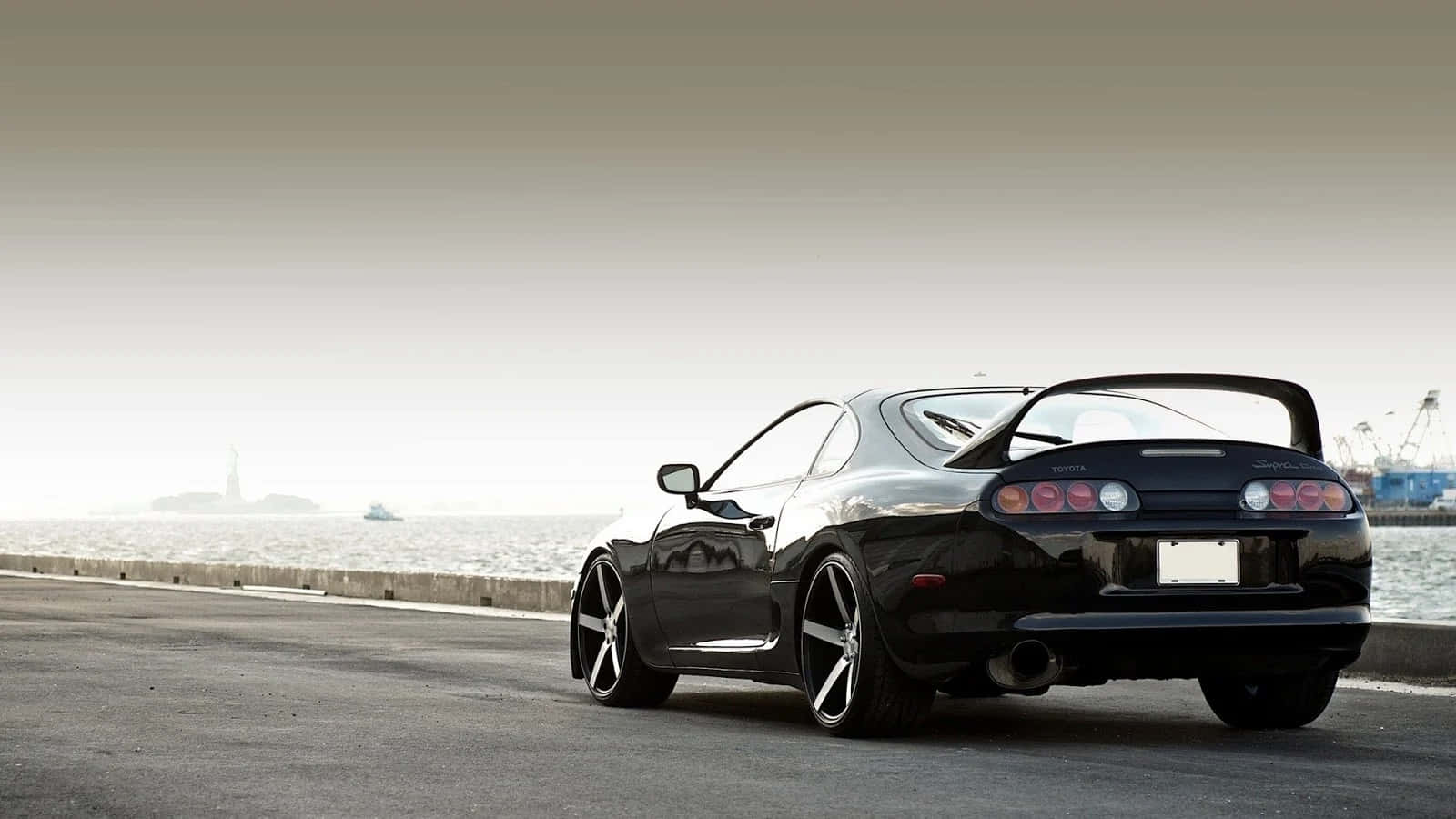 Supra 桌布 壁紙 — 一輛時尚的黑色跑車配備後擾流板，停泊在海岸公路上，俯瞰著朦朧天空下的平靜水域。