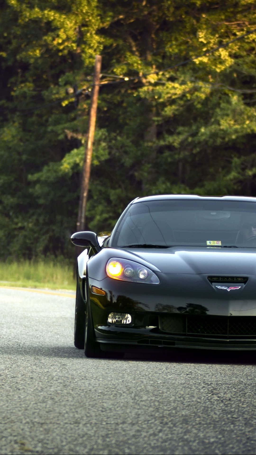 corvette 桌布 壁紙 — 一輛時尚的黑色跑車配備發光車頭燈，在黃金時段沿著綠樹成蔭的道路行駛。