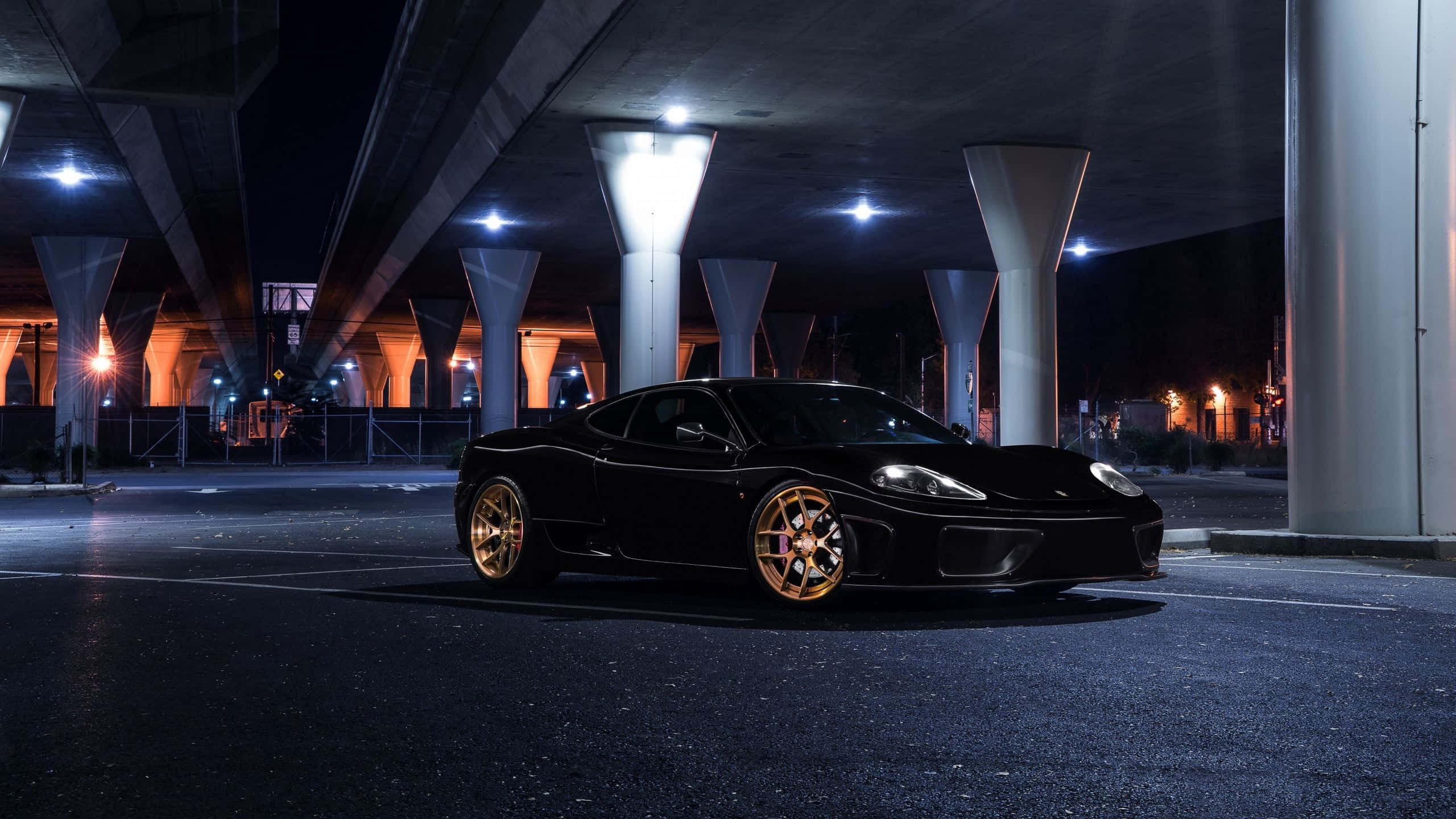 Ferrari 360 Modena 桌布 壁紙 — 一輛時尚的黑色跑車配金色輪圈，夜晚停放在燈光照亮的混凝土高架橋下。
