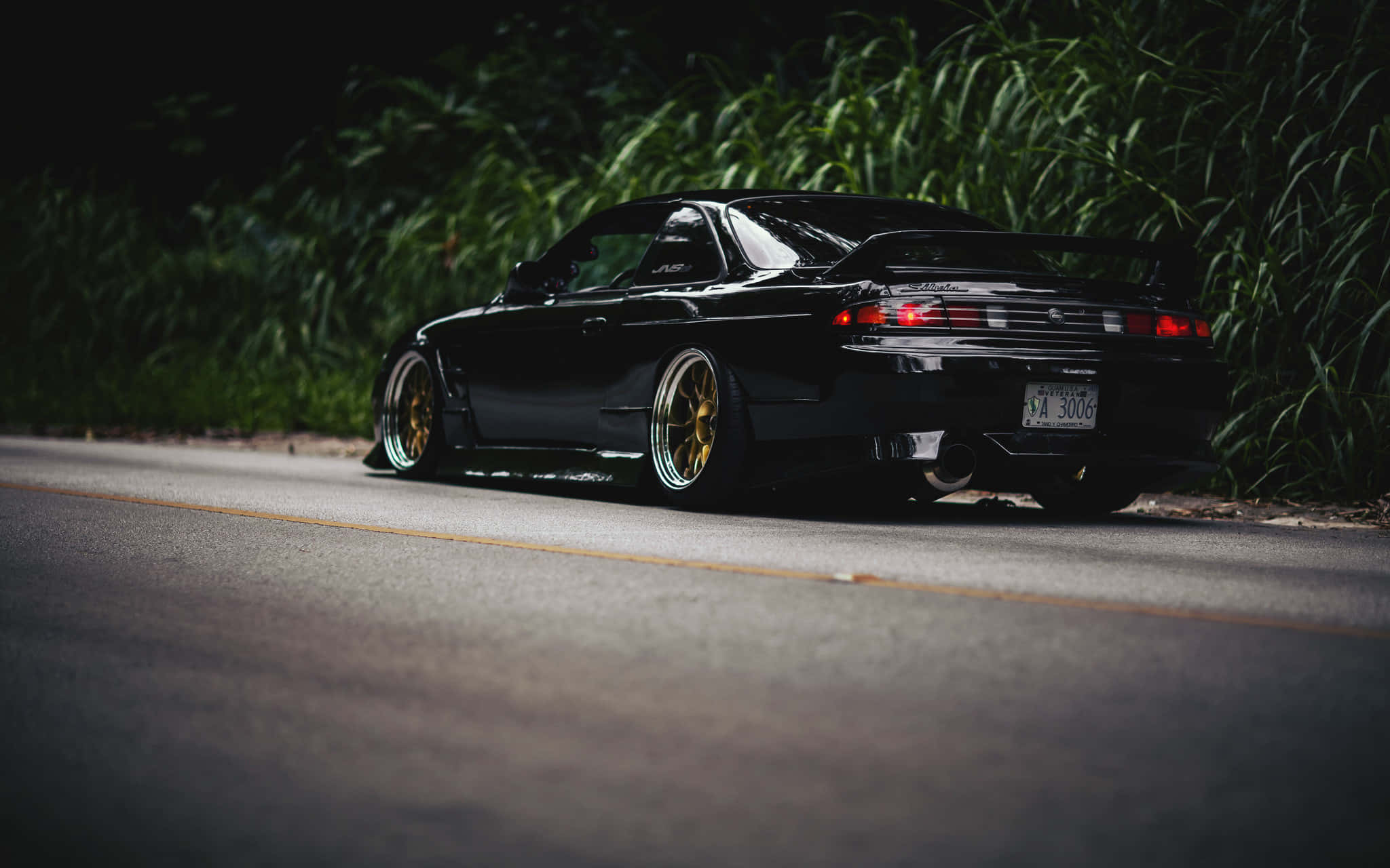 Nissan 180sx 桌布 壁紙 — 一輛時尚的黑色跑車配金色輪圈，夜間停泊在黑暗的道路上，周圍是高高的綠草。