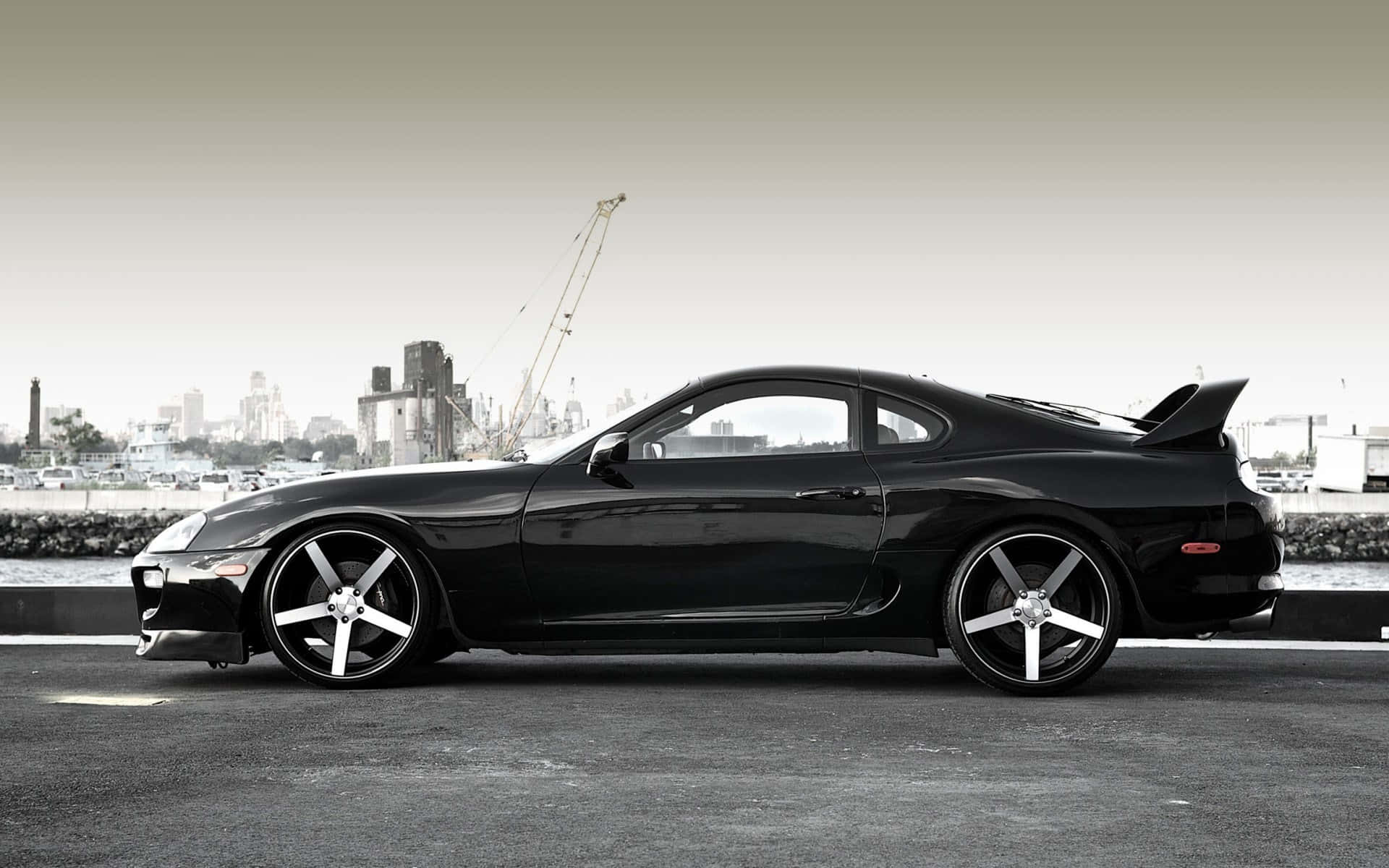 黑色跑車配銀色輪圈，停在海濱道路上，靠近城市天際線。 — Toyota Supra Mk4 壁紙