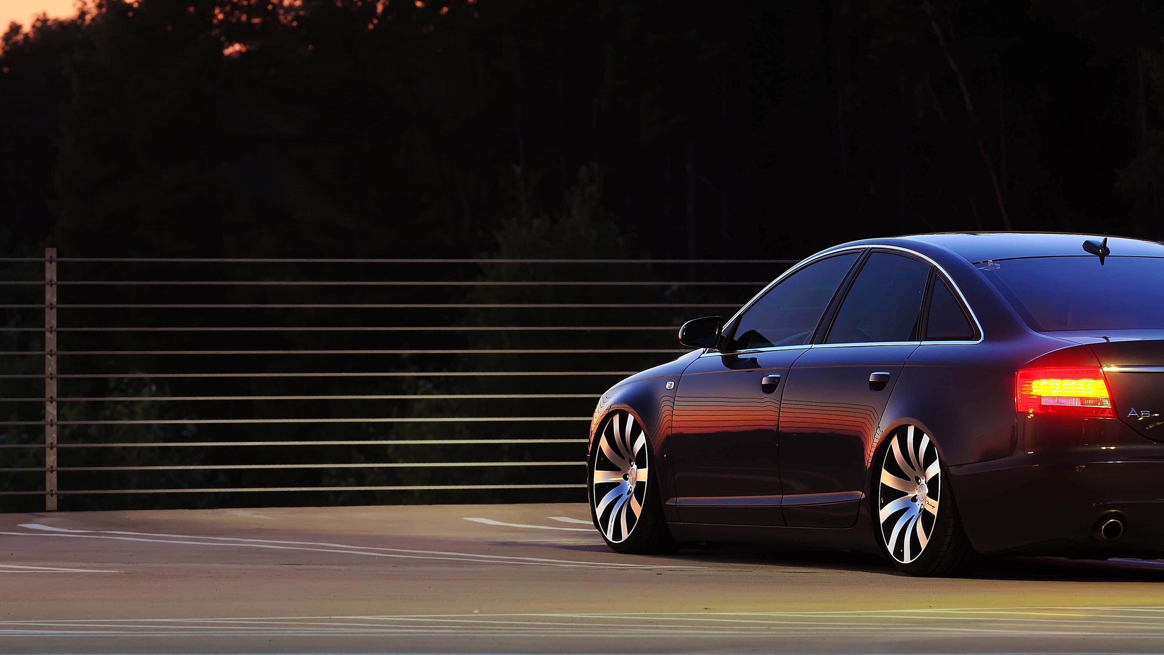 Audi A6 桌布 壁紙 — 一輛時尚的黑色轎車，帶有發光的尾燈，在日落時沿著賽道滑行，在其拋光的表面上投射出溫暖的反射。