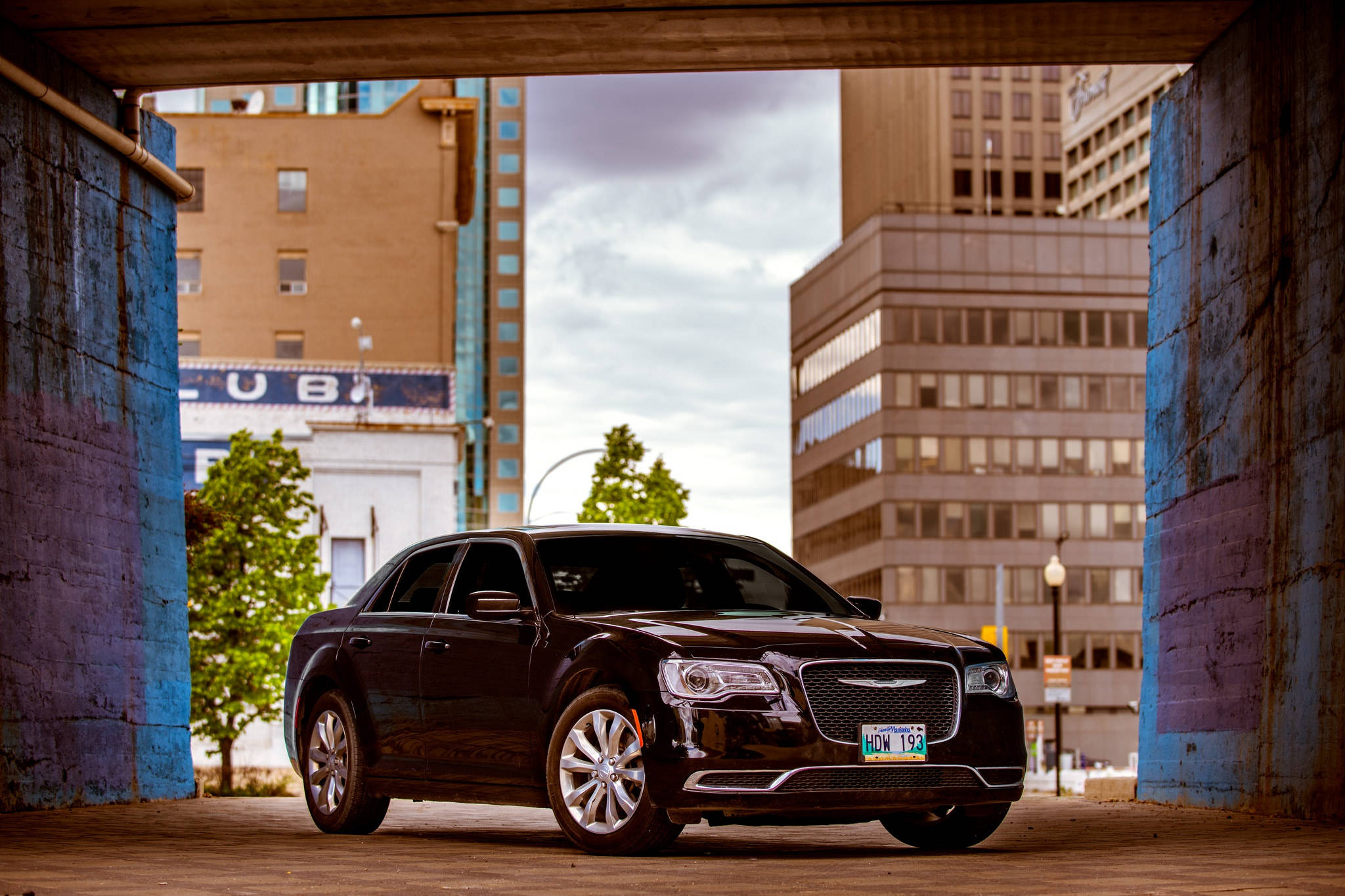 Chrysler 桌布 壁紙 — 一輛時尚的黑色轎車停泊在藍色城市高架橋下，背景有城市建築物和樹木。