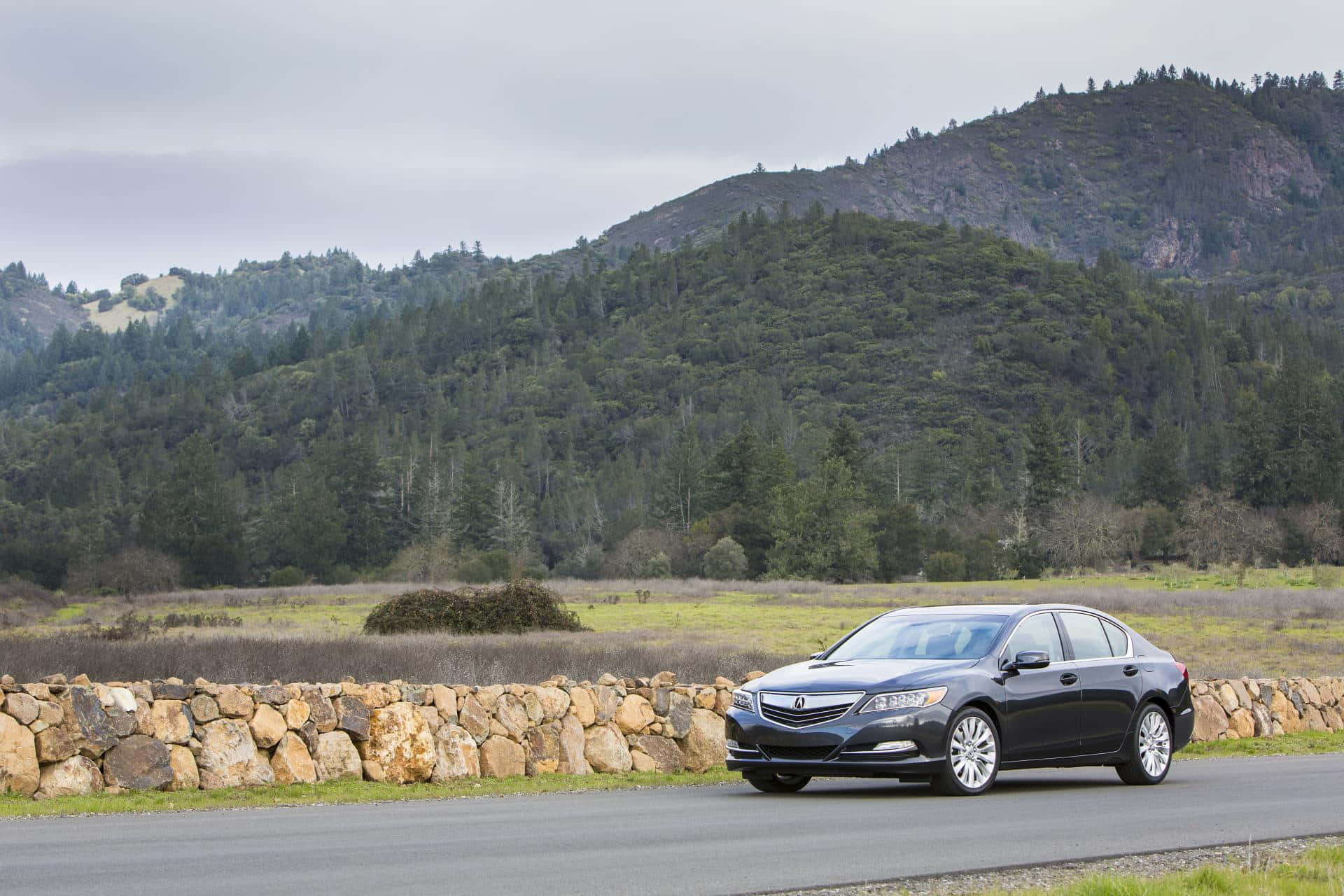 Acura Rlx 桌布 壁紙 — 一輛時尚的黑色轎車行駛在一條鋪砌的道路上，道路兩旁是石牆，後面是森林覆蓋的山丘，天空多雲。