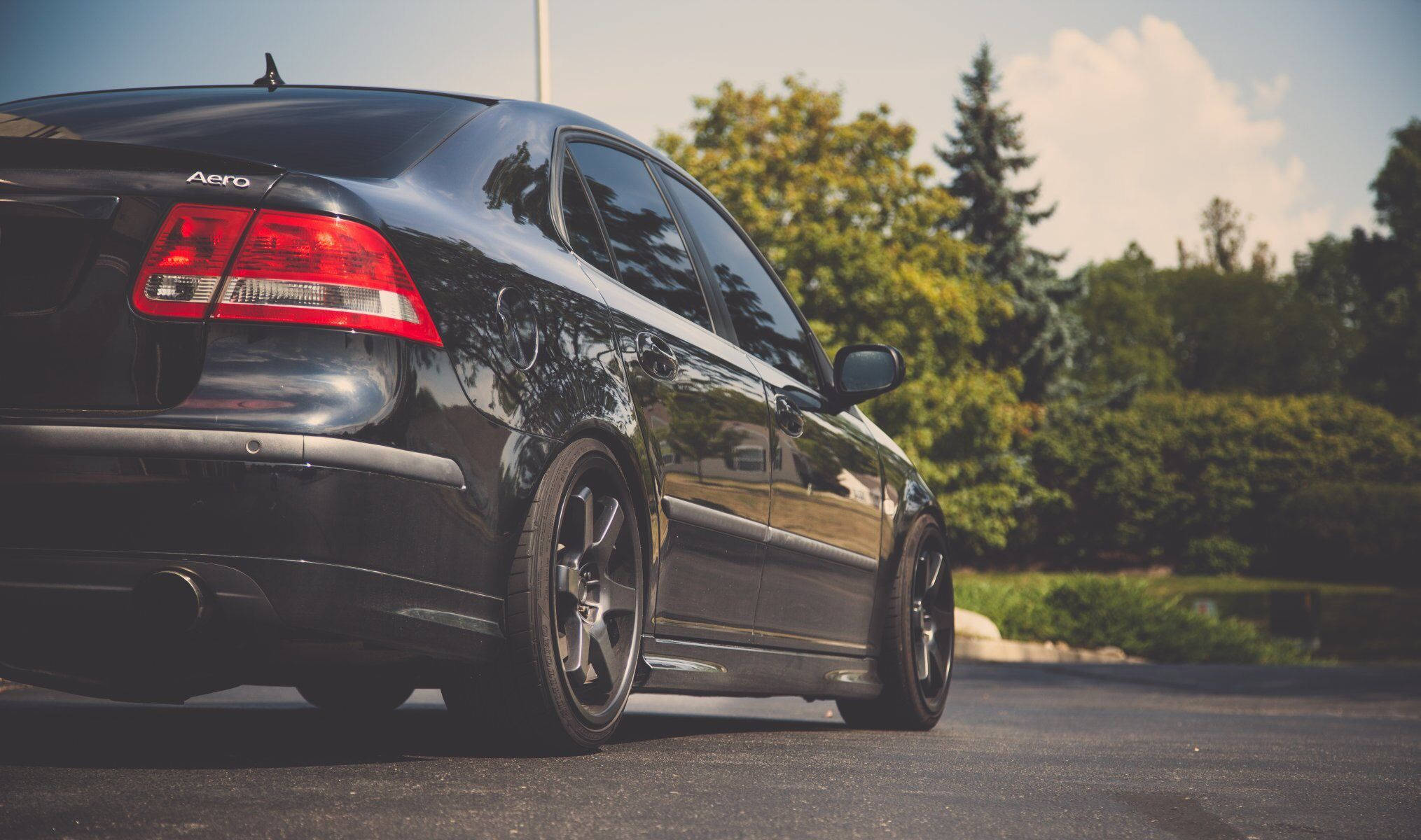 Saab 桌布 壁紙 — 一輛時尚的黑色轎車配備定制輪圈，停在瀝青路面上，背景是明亮的天空和綠樹。