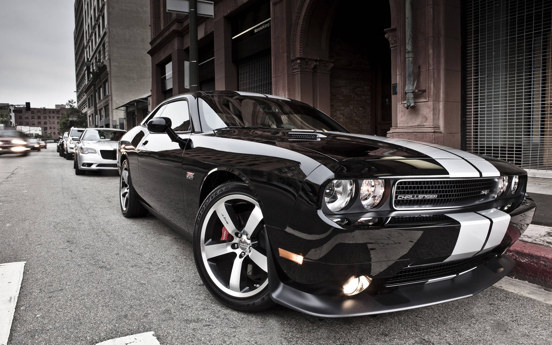 dodge iPhone 桌布 壁紙 — 一輛時尚的黑色道奇挑戰者，帶有白色賽車條紋，停泊在歷史磚砌建築旁邊的城市街道上。