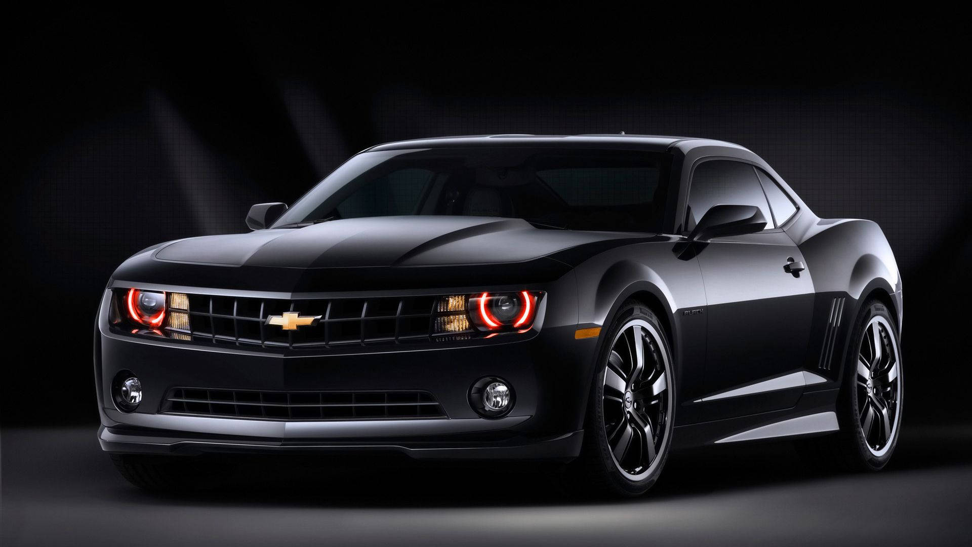Chevrolet 桌布 壁紙 — 一輛時尚的黑色雪佛蘭 Camaro，帶有發光的紅色頭燈，在黑暗的攝影棚背景下戲劇性地照亮。