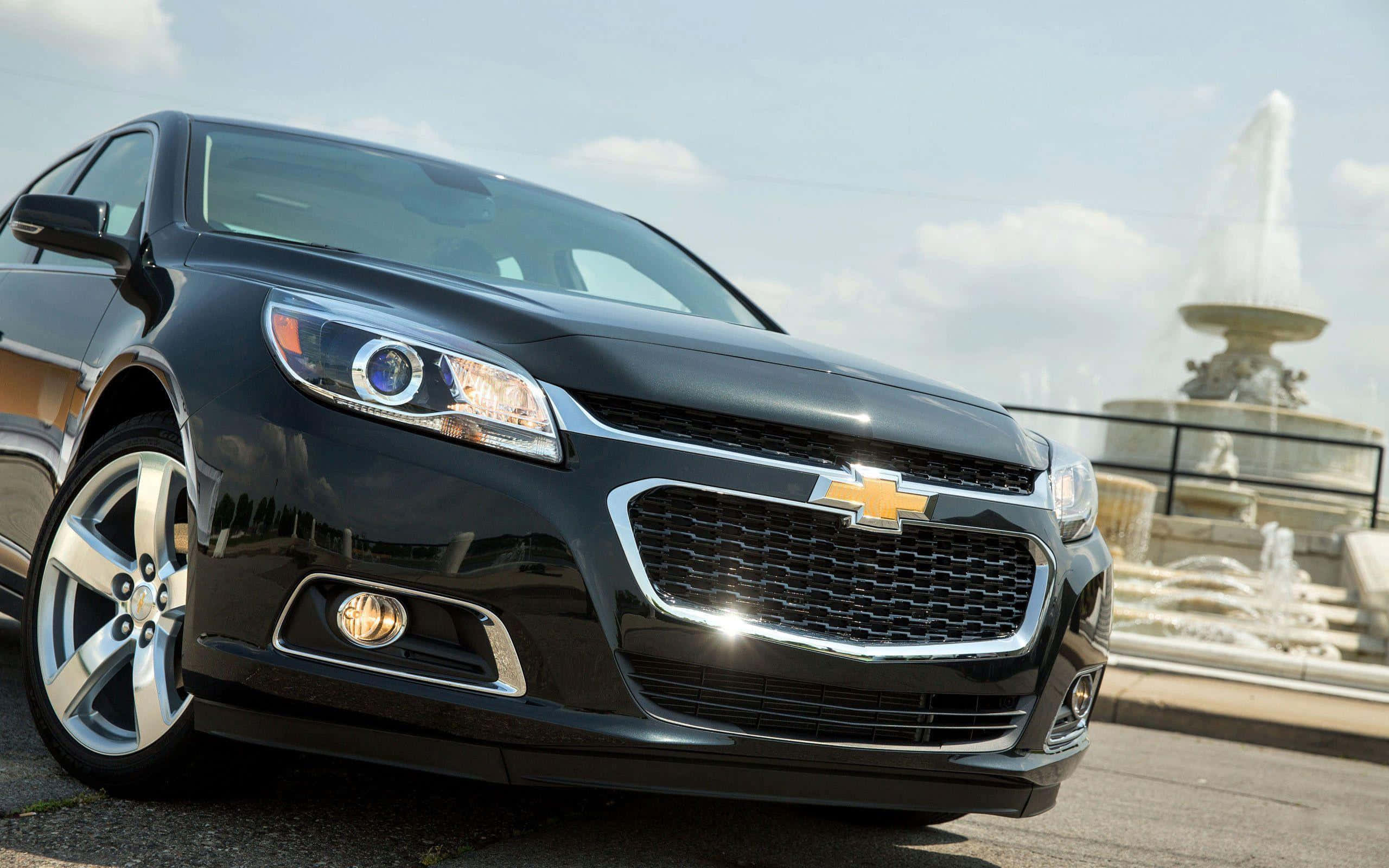 chevy 桌布 壁紙 — 一輛時尚的黑色雪佛蘭轎車在明亮的日光下閃閃發光，背景是宏偉的噴泉。