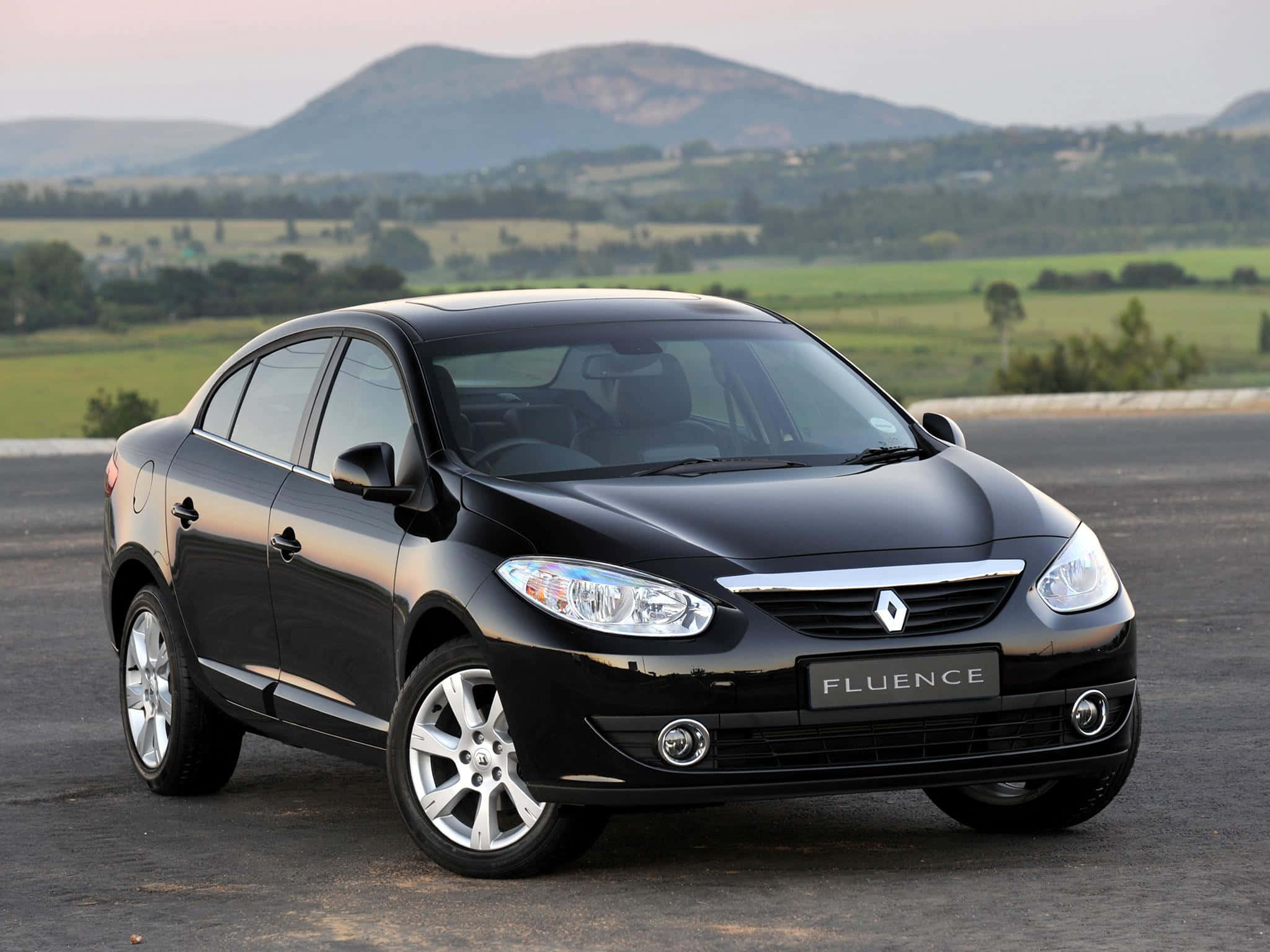 Renault Fluence 桌布 壁紙 — 一輛時尚的黑色雷諾 Fluence 轎車停在柏油路上，背景是起伏的綠色丘陵和遠山，天空呈現柔和的暮色。