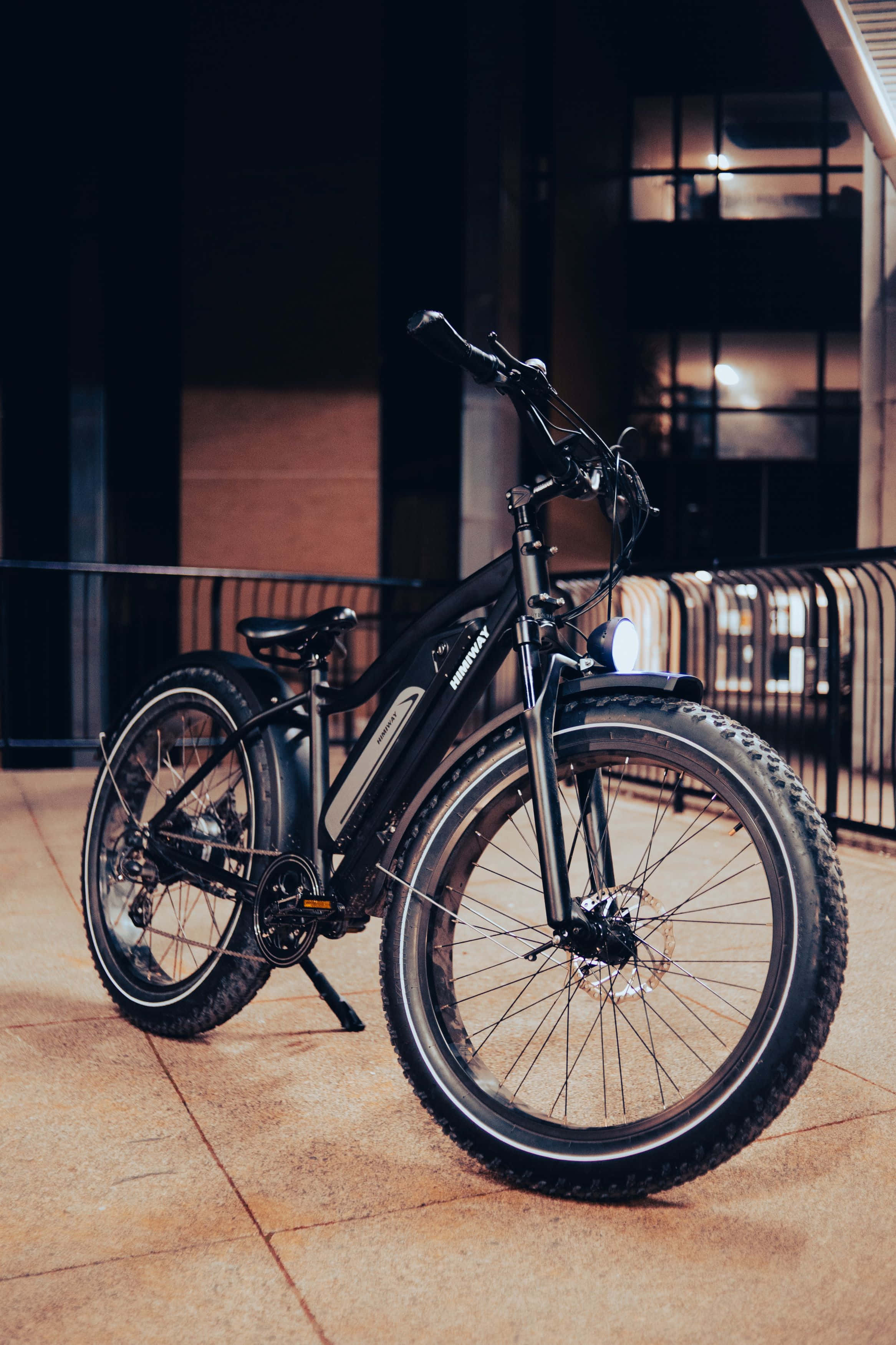 Electric Bike 桌布 壁紙 — 一輛時尚的黑色電動自行車配備厚輪胎，夜晚停放在磁磚人行道上，附近有一座現代建築，室內燈光溫暖。