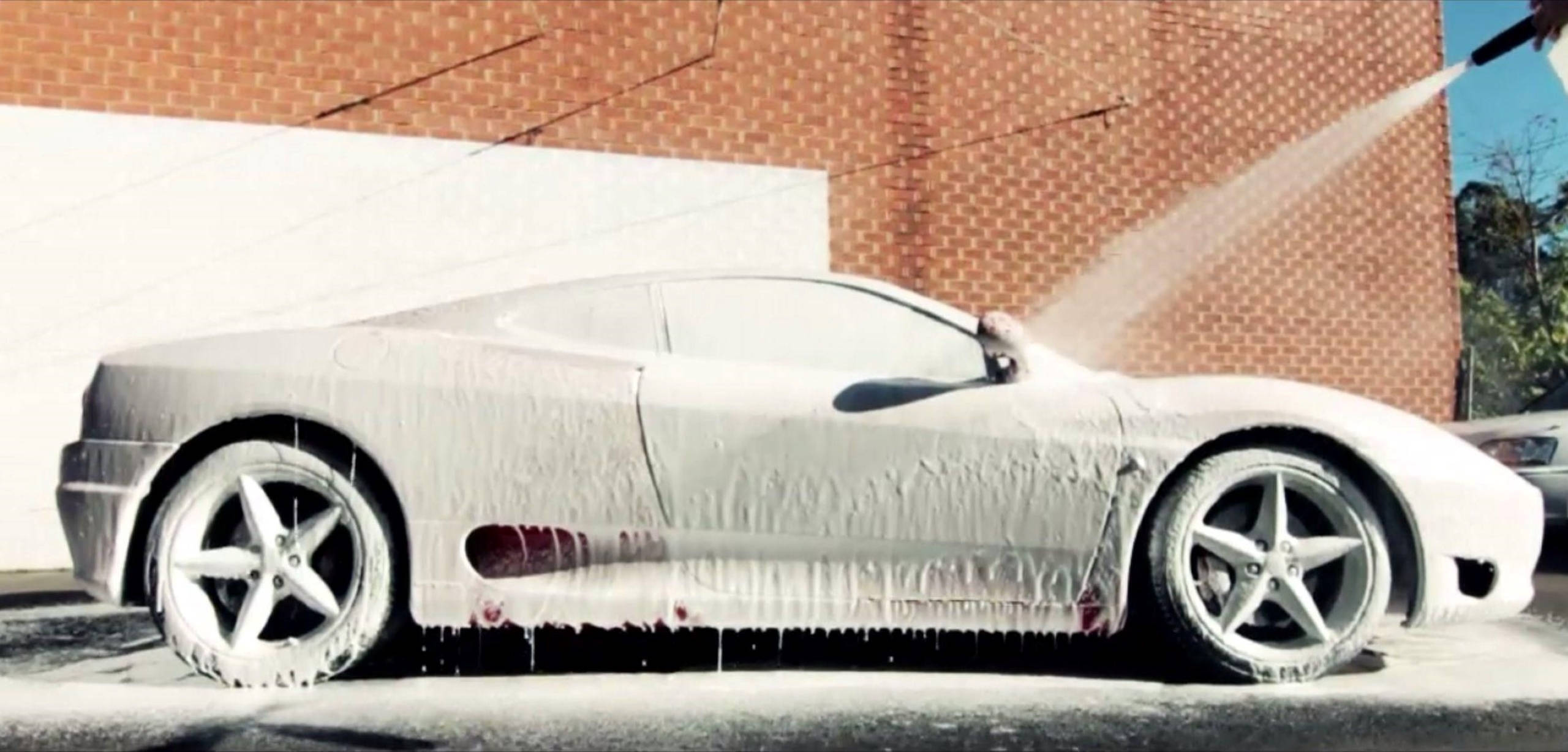 Wash 桌布 壁紙 — 一輛時尚跑車覆蓋著厚厚的白色肥皂泡，在明亮的陽光下正對著磚牆進行高壓清洗。